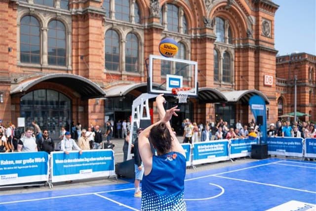 Eine Basketballspielerin wirft von der 3-Punkte-Linie. Im Hintergrund sind Zuschauer und der Bremer Hbf zu sehen.