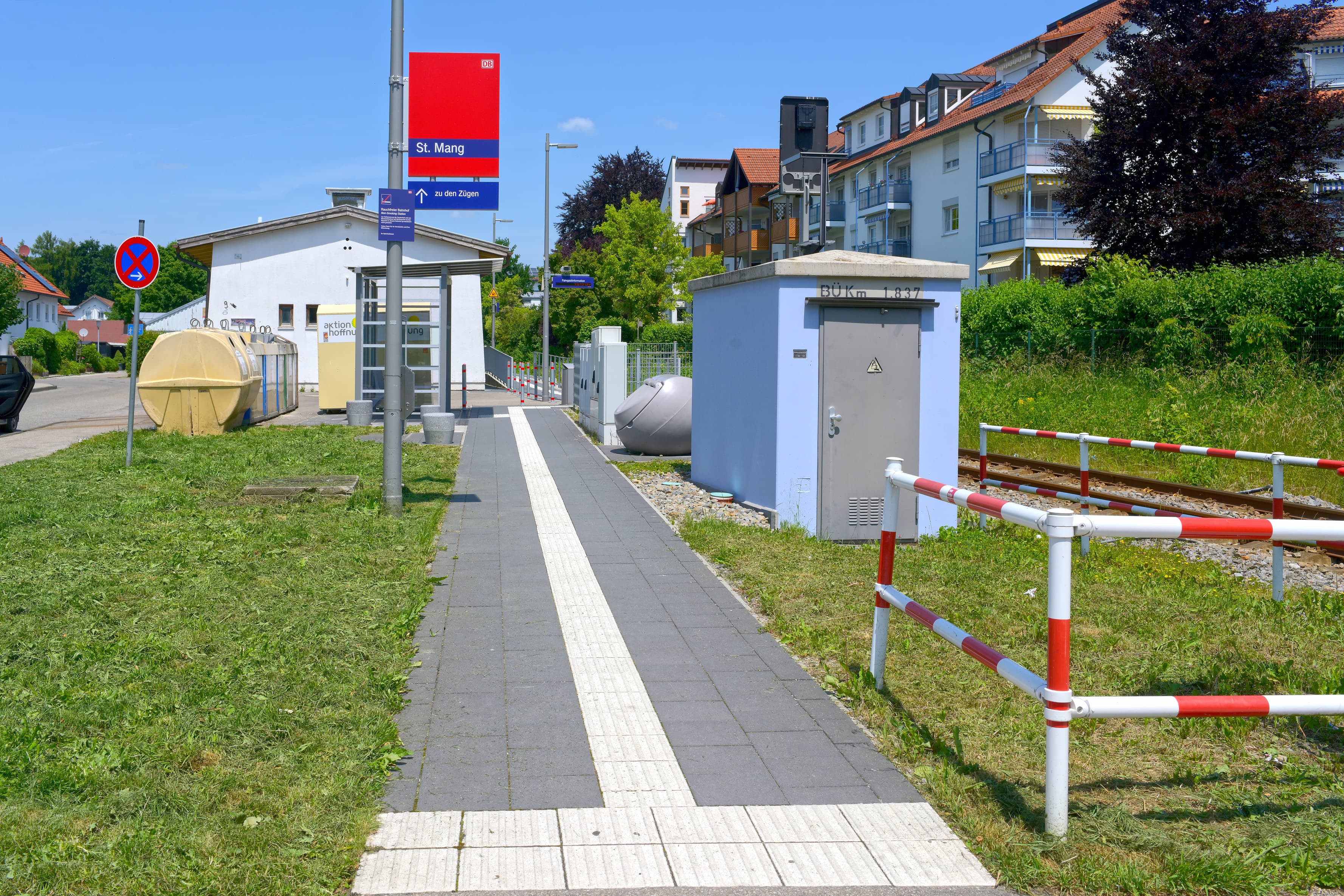 Zentral im Bild die stufenlose Zuwegung zum Bahnsteig mit taktilem Leitsystem. Hinten links im Bild ein modernes Wetterschutzhaus. Vorne rechts ein blau gestrichenes Gerätehäuschen vor den Gleisen.