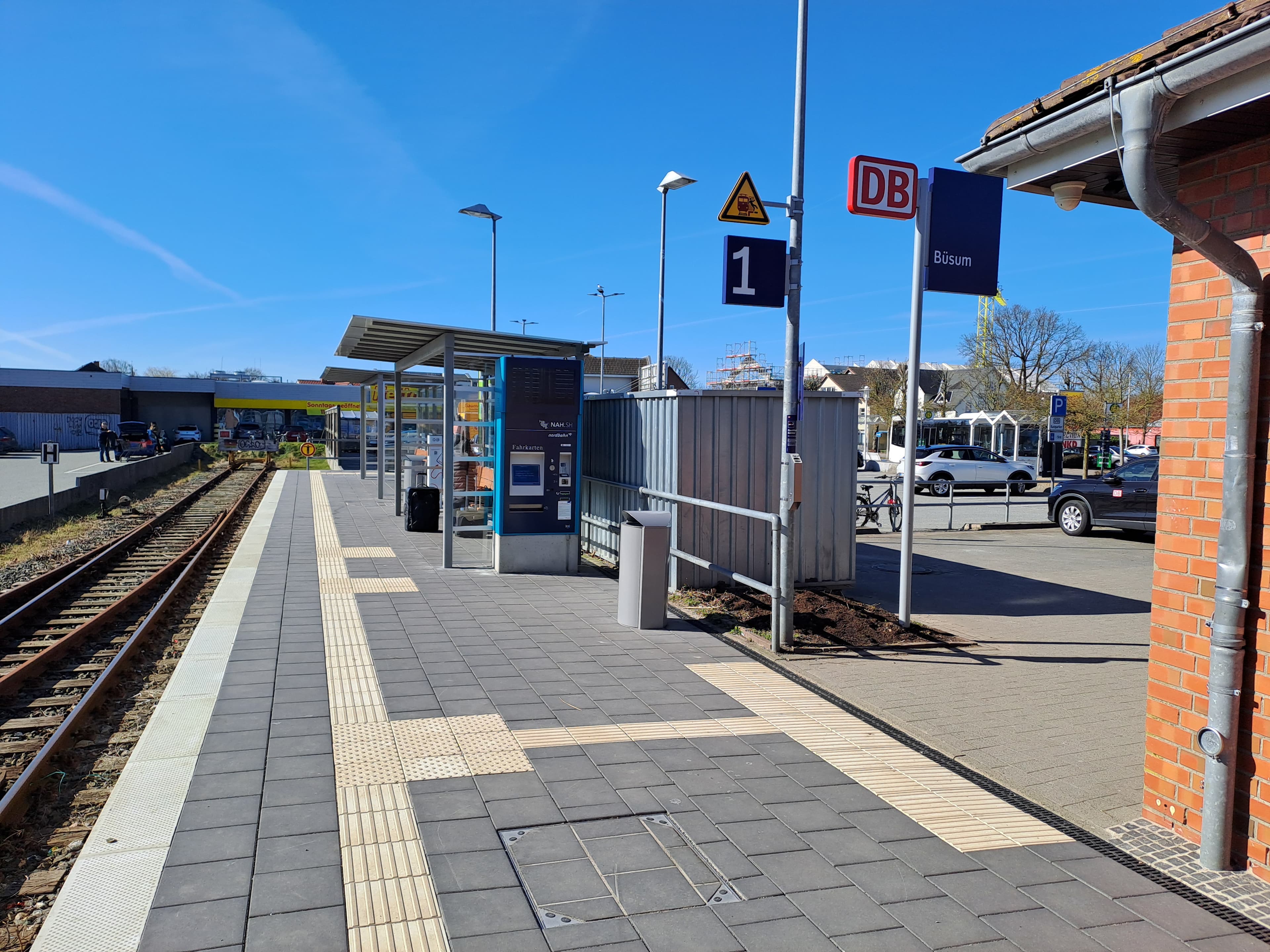 Im Bild der Bahnsteig mit neuem Belag und Blindenleitstreifen, zwei Wetteschutzhäusern mit Holzsitzbänken und der neuen Bahnhofsfahne am Bahnsteigzugang.
