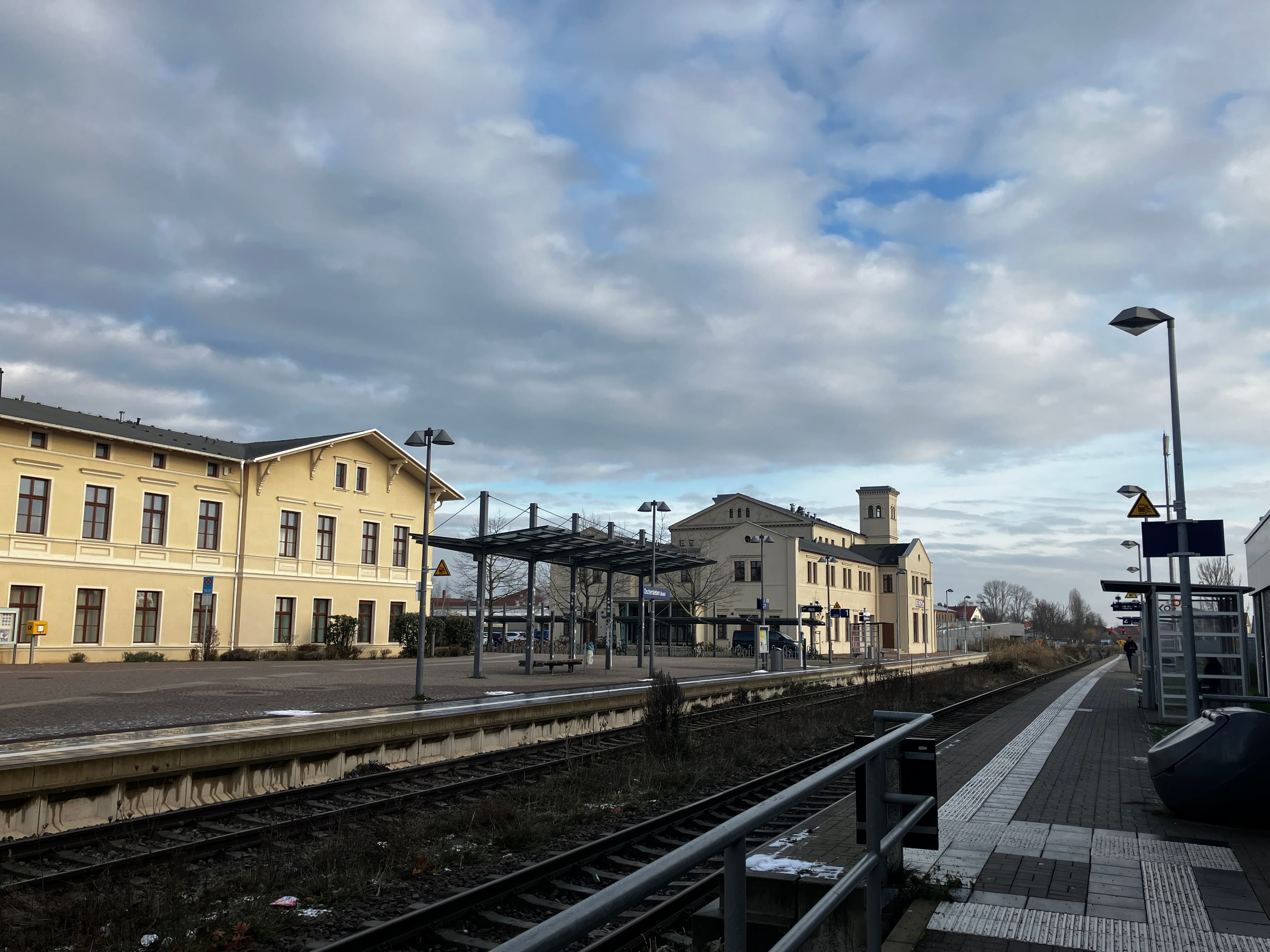 Zweistöckiges Empfangsgebäude mit hellgelber Fassade im Hintergrund, im Vordergrund beide Bahnsteige und ein Glasdach