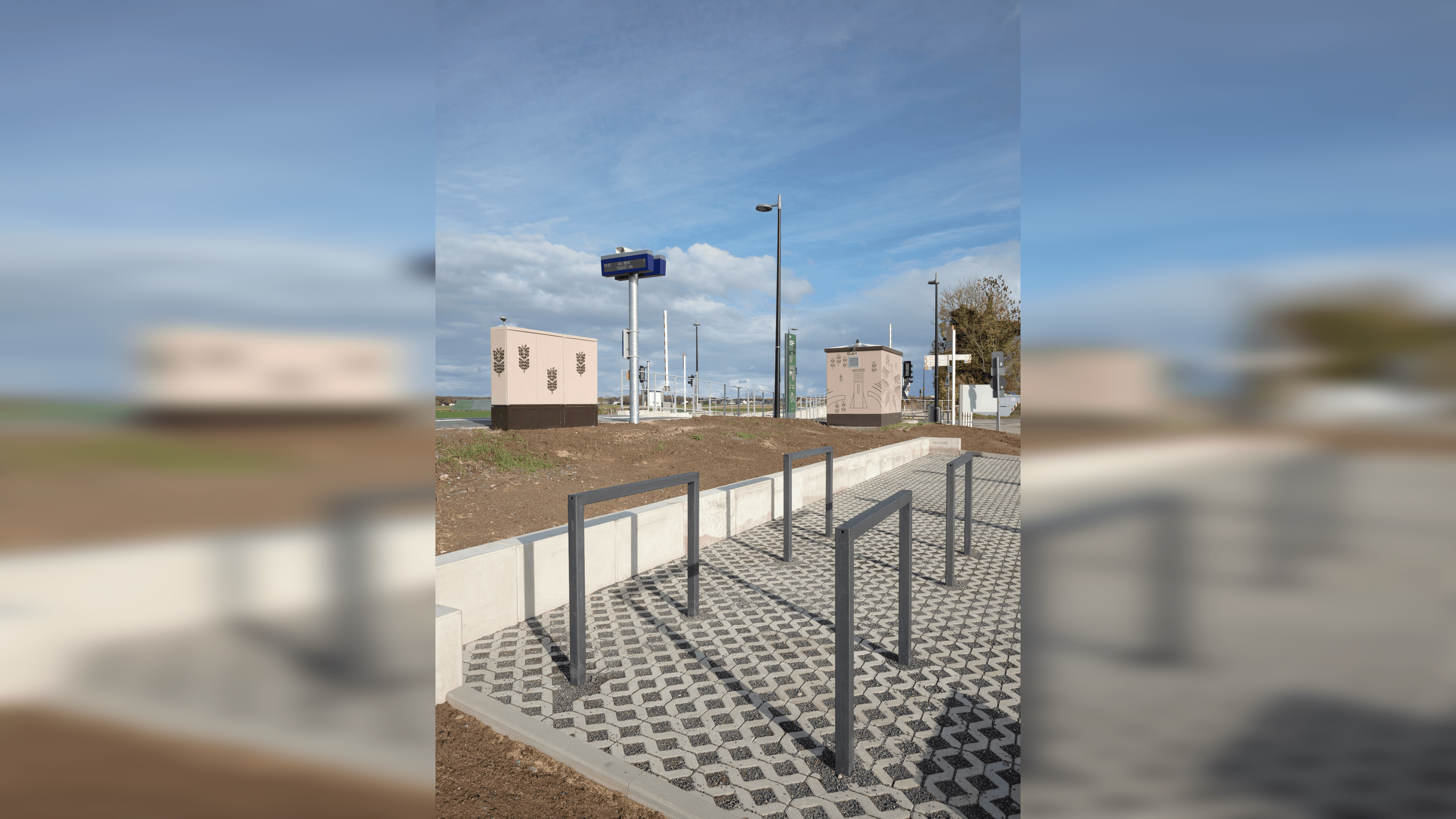 Vier Fahrradbügel in dunkelgrauer Farbe in einer gepflasterten Einbruchtung neben dem Bahnsteig. Das Technikhäuschen und der Dynamische Schriftenanzeiger sind auf dem Bahnsteig.