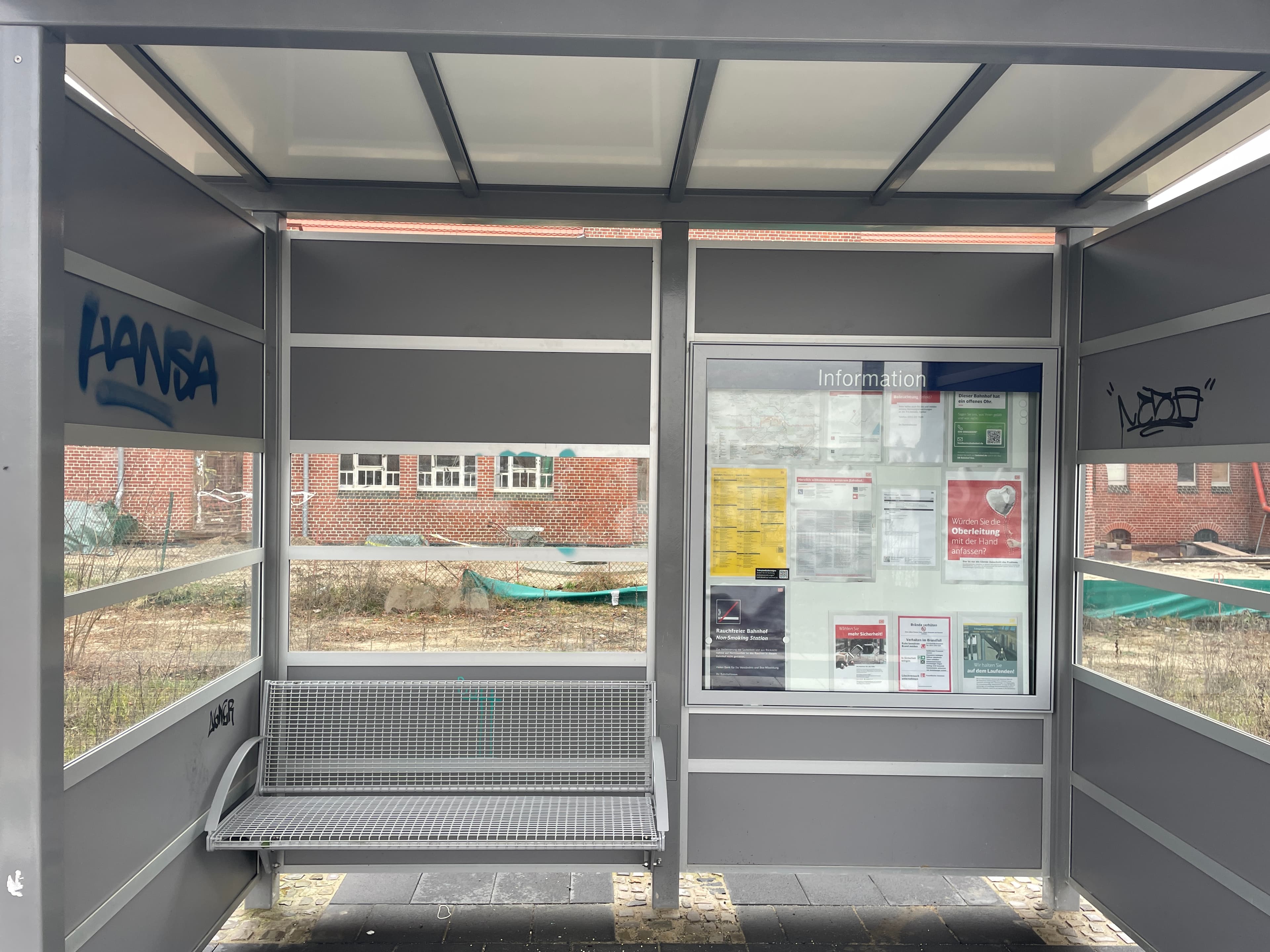 Blick in ein graues Wetterschutzhäuschen mit Sitzbank und Infovitrine.