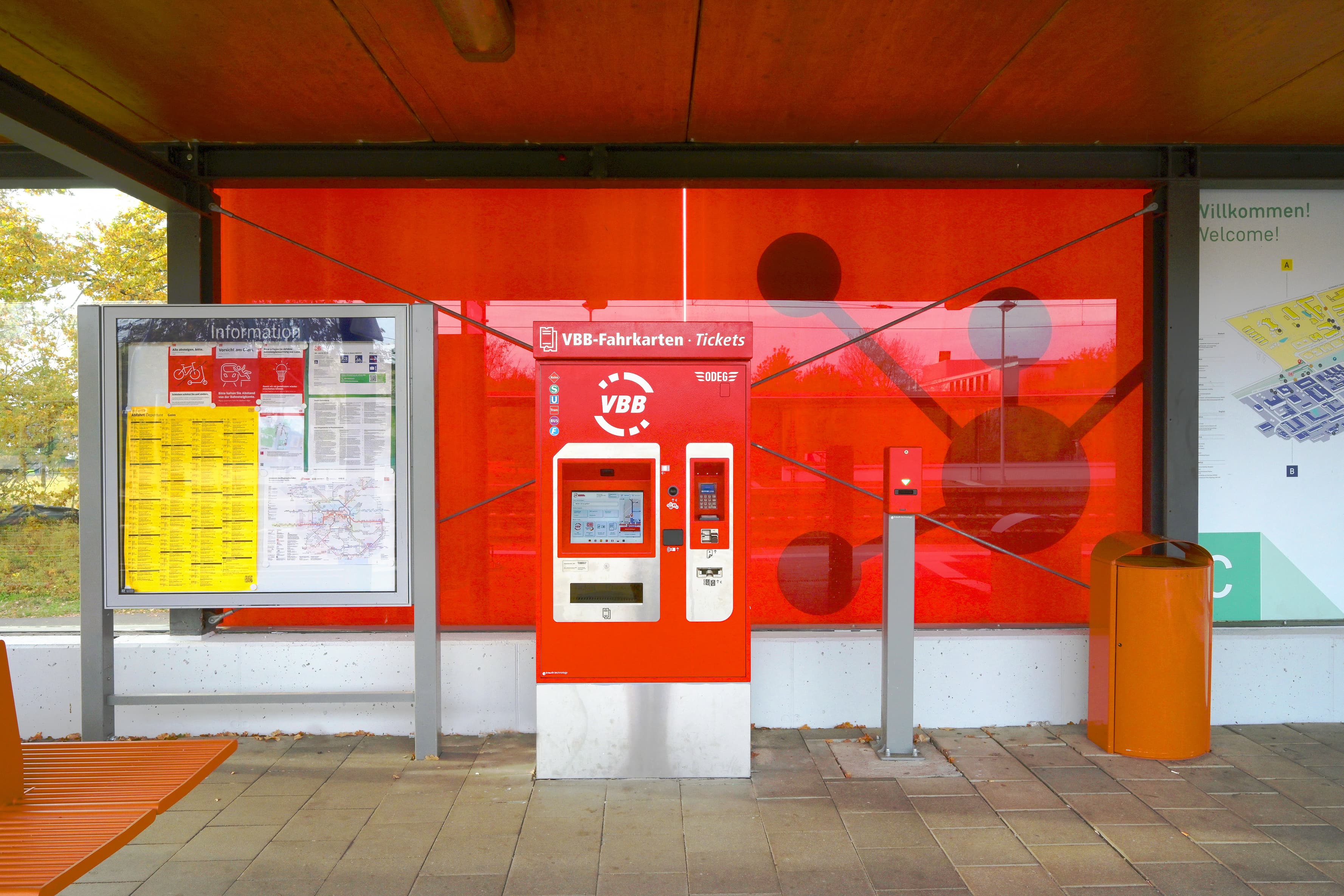 Blick auf den erneuerten Wartebereich. Zu sehen sind die neue Vitrine, ein neuer Automat und ein neuer Abfallbehälter (vlnr). Dahinter ist nun eine große, rote Glasfläche, durch die schönes Licht auf den Bahnsteig fällt.