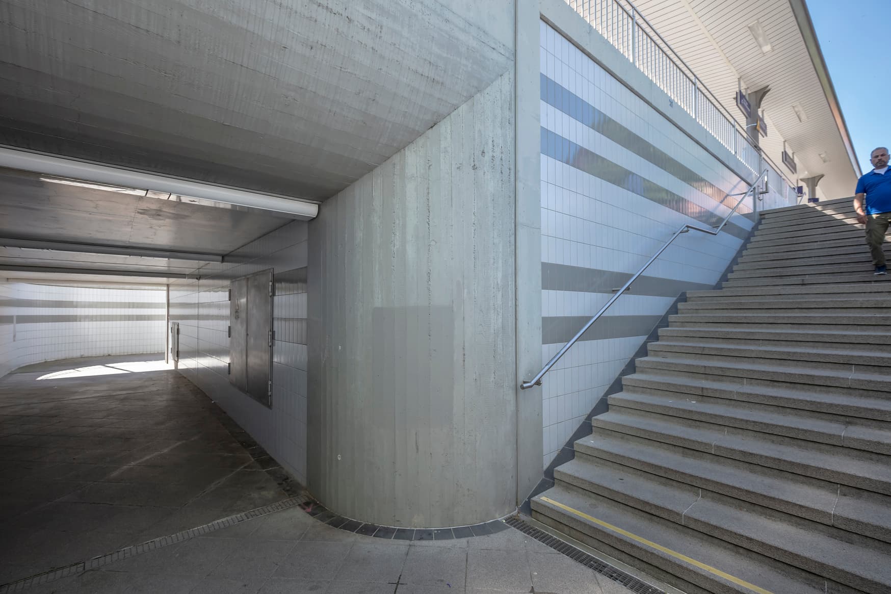 Blick auf eine Unterführung. Rechts und links entlang der Treppe befindet sich ein Handlauf. Die Wände der Unterführung sind weiß/grau gefliest.