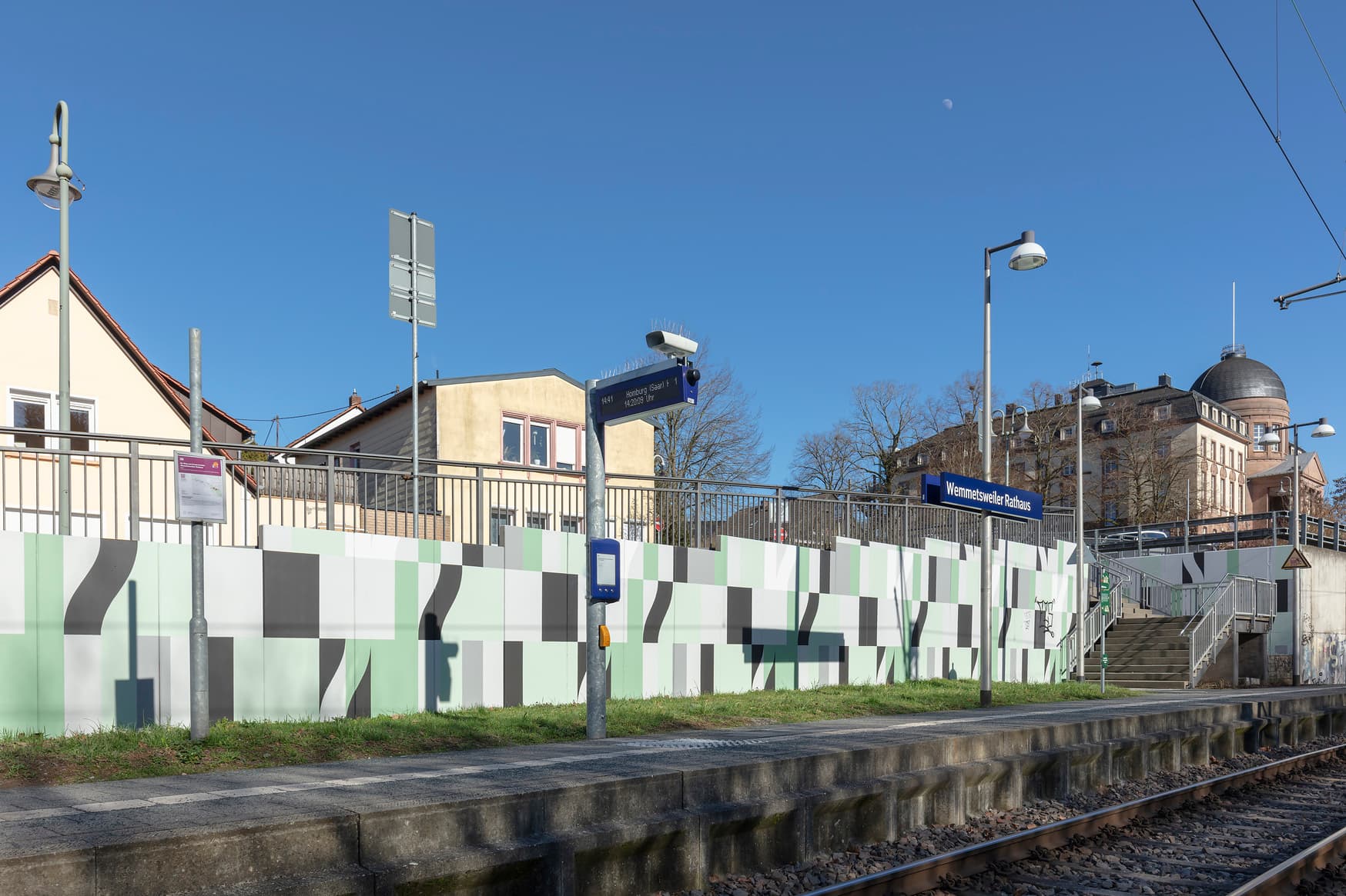 Blick auf einen Bahnsteig. Davor befindet sich das Gleis. Im Hintergrund eine bunt geflieste Mauer und rechts eine Treppe.