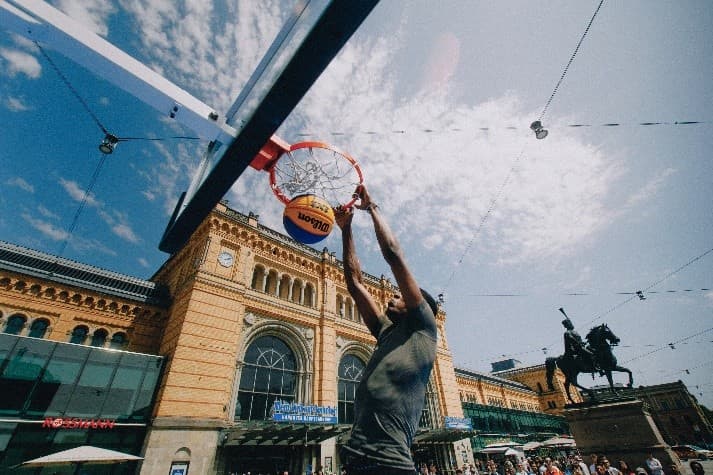 Ein Basketballspieler dunkt vor dem historischen Bahnhofsgebäude des Hannover Hbf im Freien, während Menschen zuschauen und eine Reiterstatue im Hintergrund zu sehen ist.
