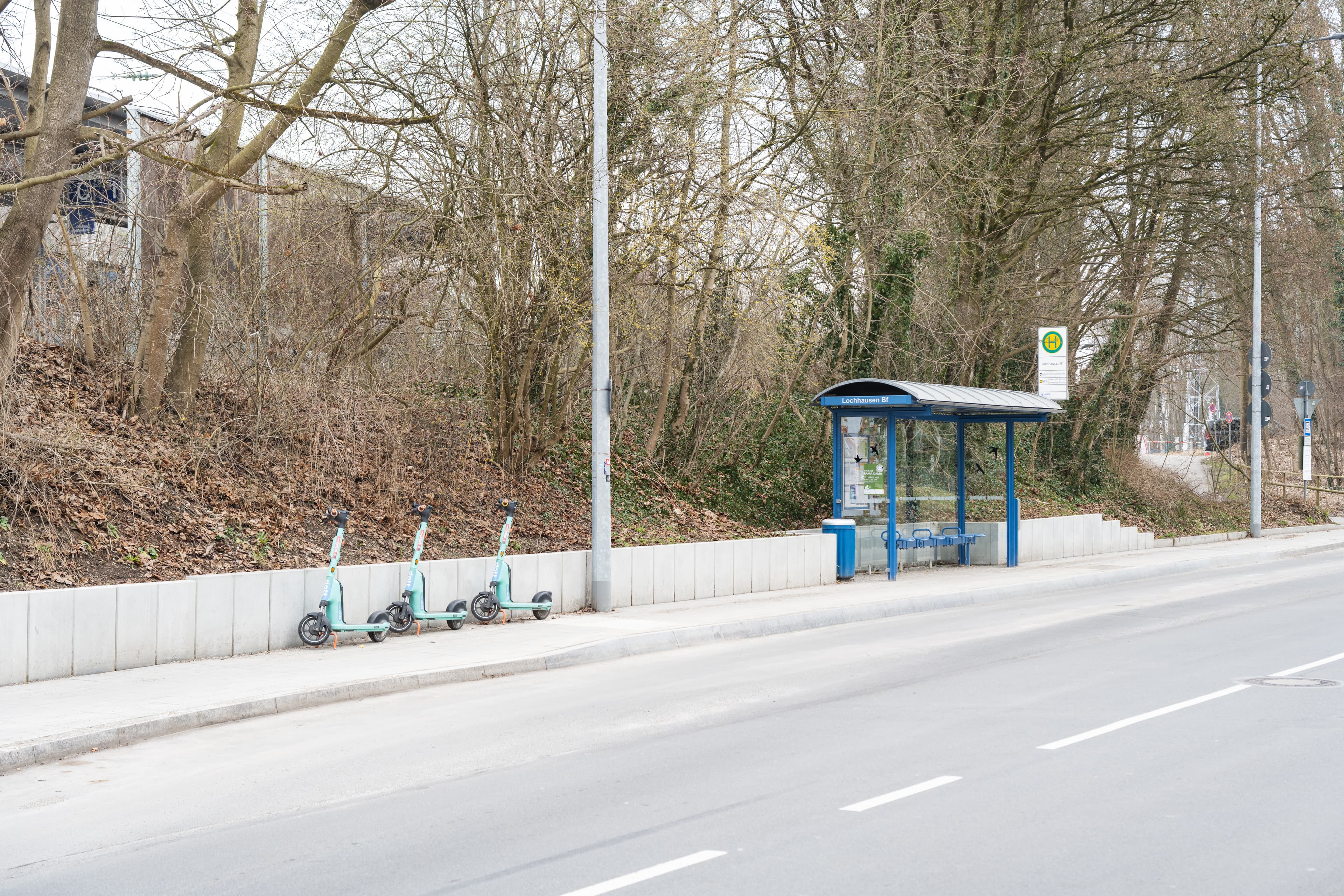 Blick auf eine Straße. Auf dem Gehweg dahinter befindet sich eine Bushaltestelle. Links davon sind drei E-Roller geparkt.