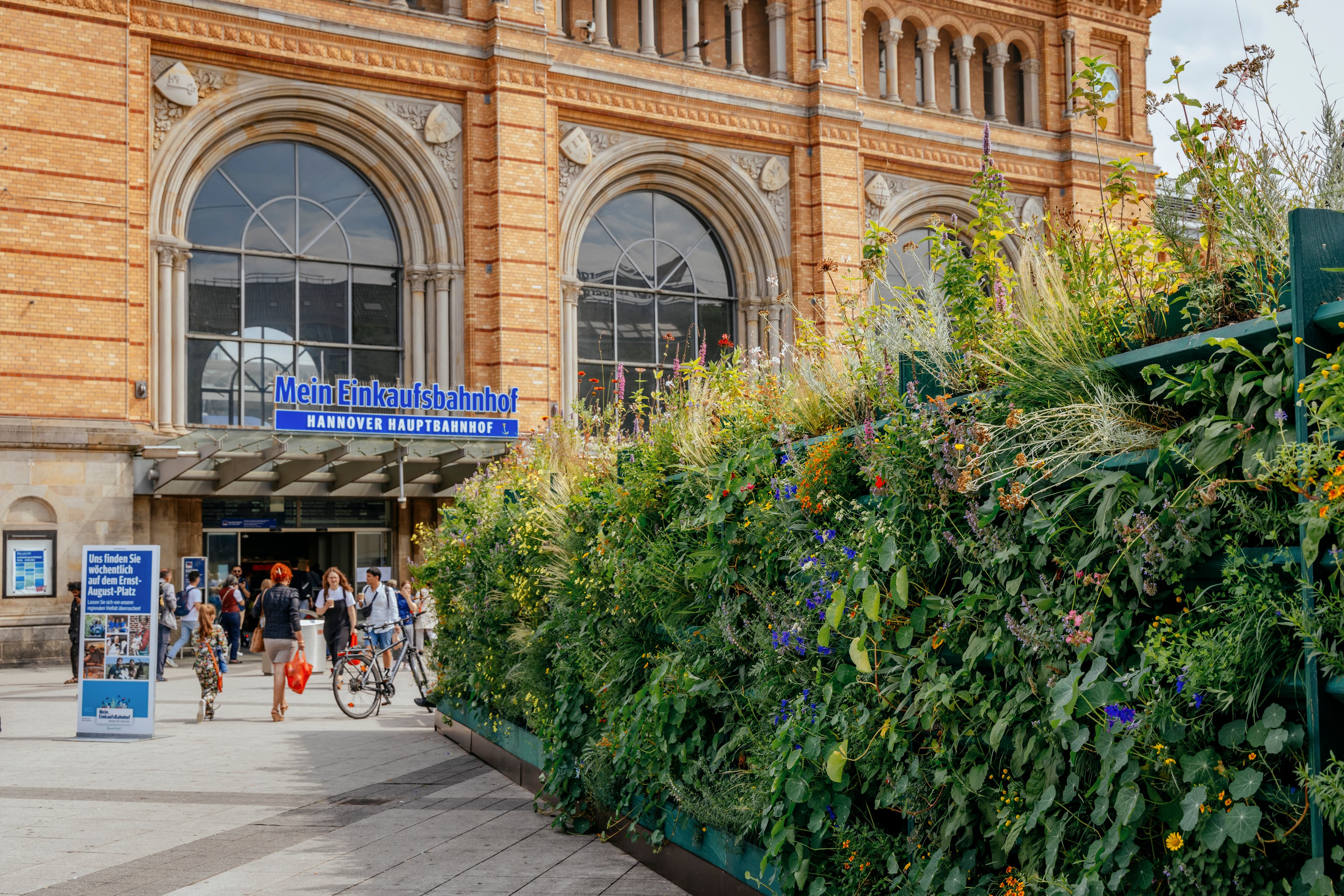 Blick auf die bepflanzte Rückwand des hölzernen Einkaufsbahnhof-Schriftzugs. Im Hintergrund ist das historische Bahnhofsgebäude des Hannover Hbf und einige Passant:innen zu sehen. 