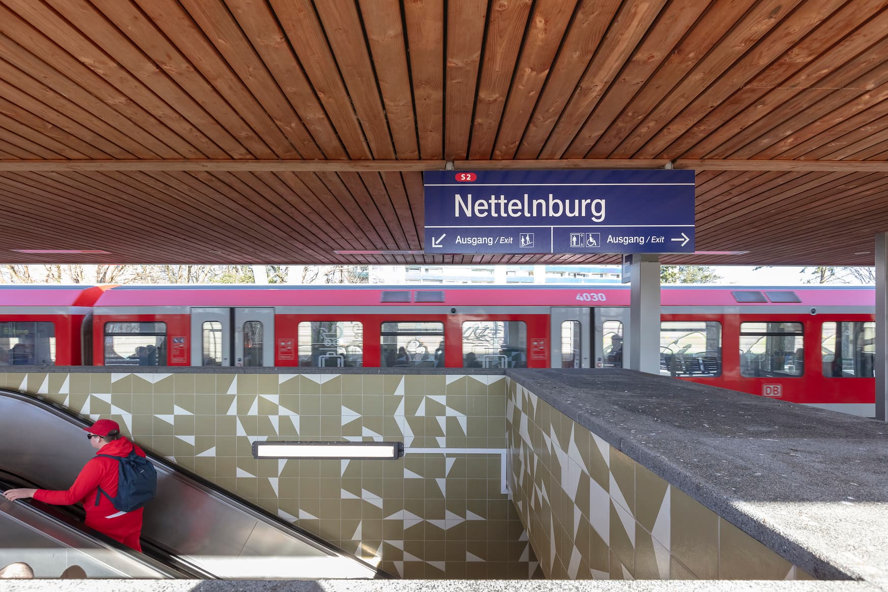 Treppenaufgang zum Bahnsteig mit grün-weißem Fliesenmuster. Ein roter Zug steht im Hintergrund, davor das Bahnhofsschild mit Wegeleitung erkennbar.