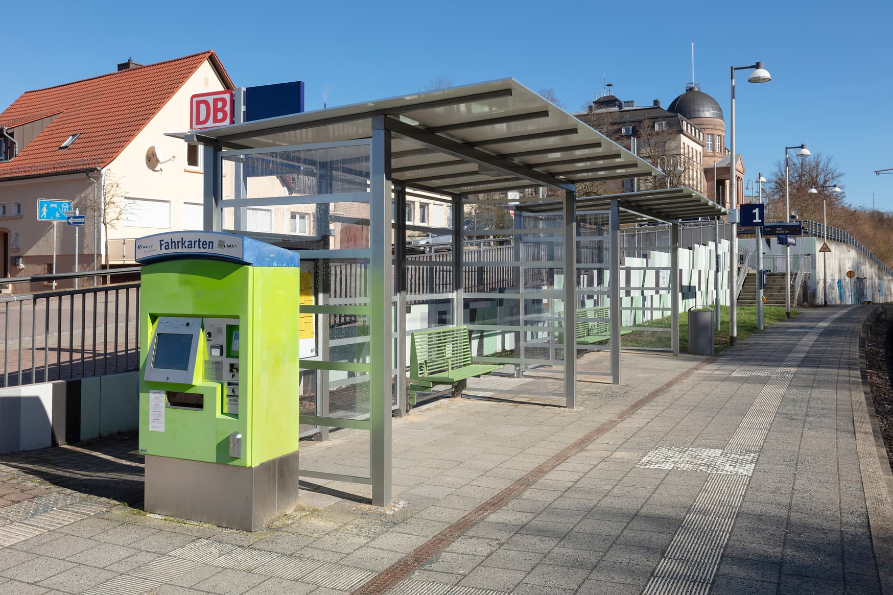 Rechts befinden sich Gleise, links davon Sitzgelegenheiten unter einem Wetterschutzhäuschen. Dazwischen der Bahnsteig.