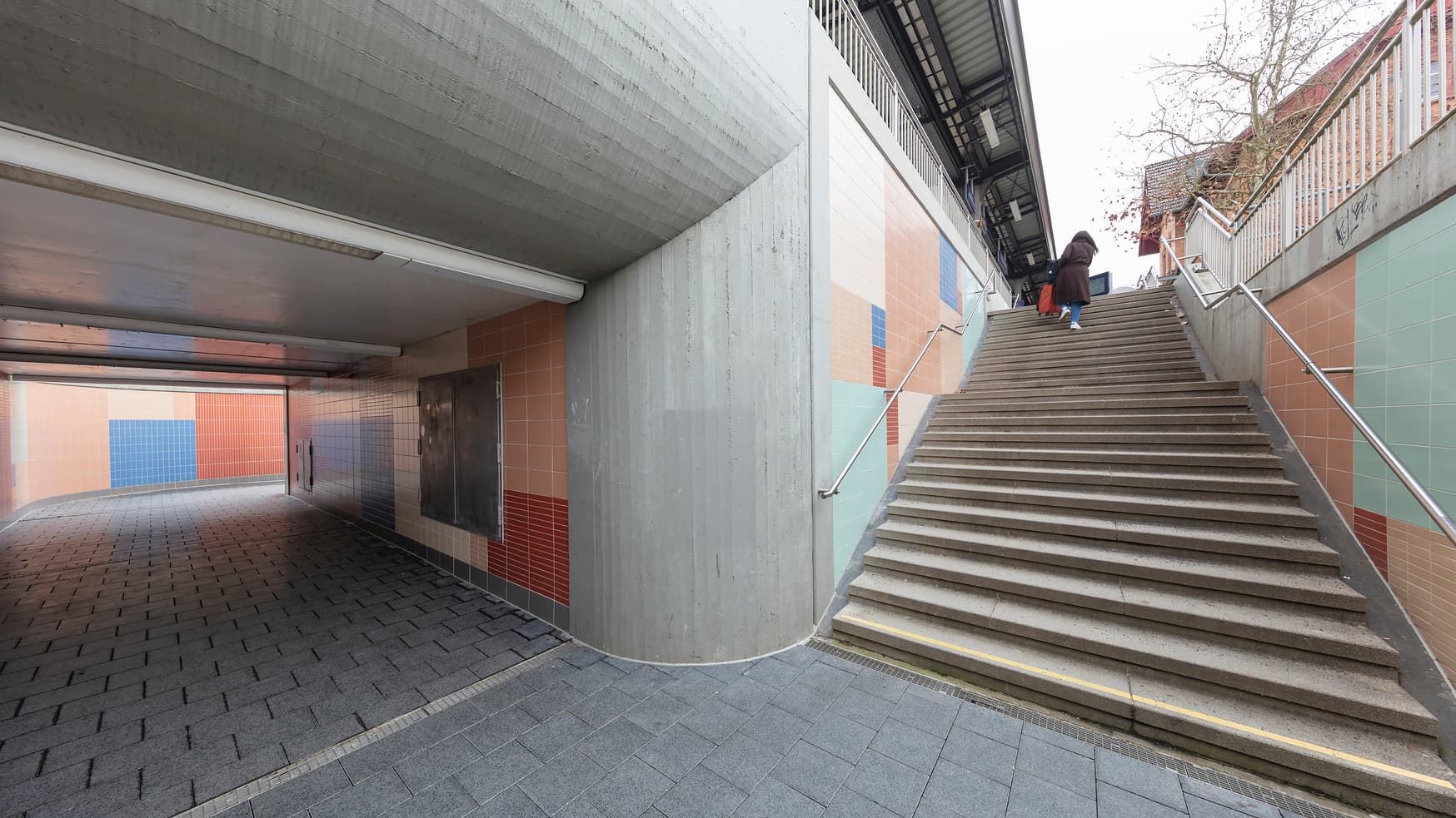 Blick auf eine Unterführung. Rechts und links entlang der Treppe befindet sich ein Handlauf. Die Wände der Unterführung sind bunt gefliest.