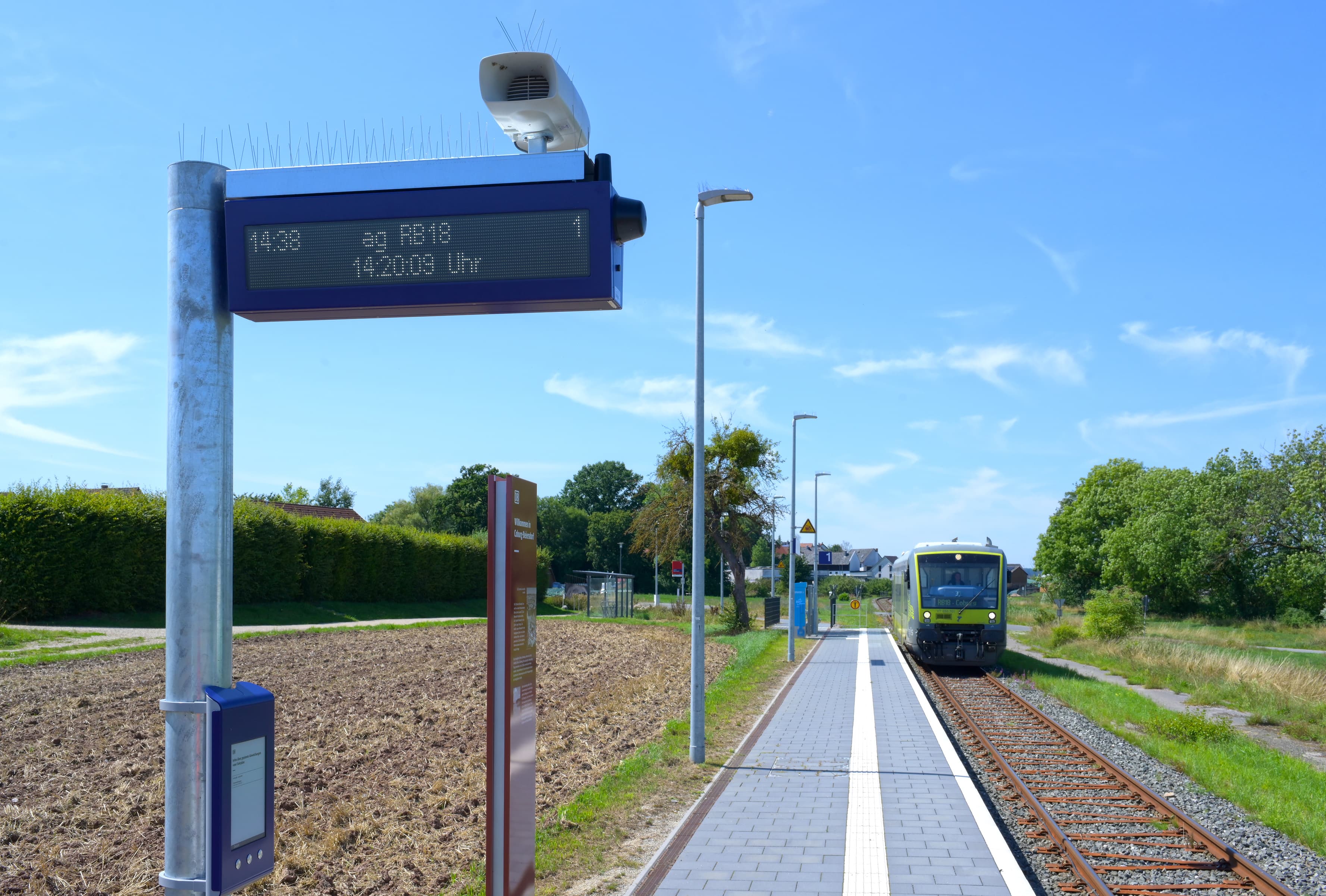 Im Vordergrund auf Brusthöhe ein DIN A4 formatiger, digitaler Aushang an einem runden Metallpfosten. Darüber ein zweizeiliger digitaler Abfahrtszeitenanzeiger mit einem Lautsprecher obenauf. 