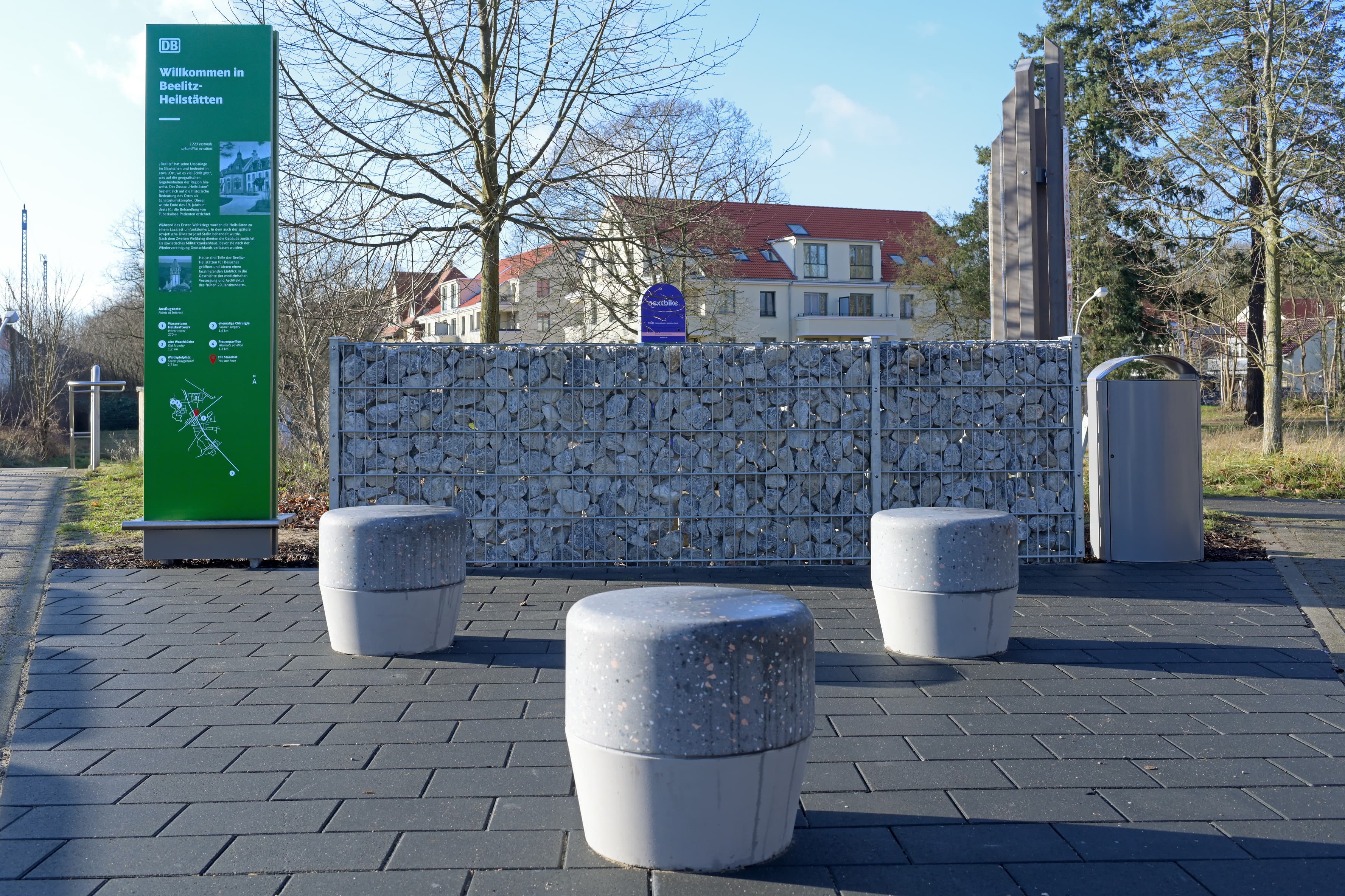 Drei Hocker aus Stein stehen vor einer Steinwand, links steht eine Informationsstele links.