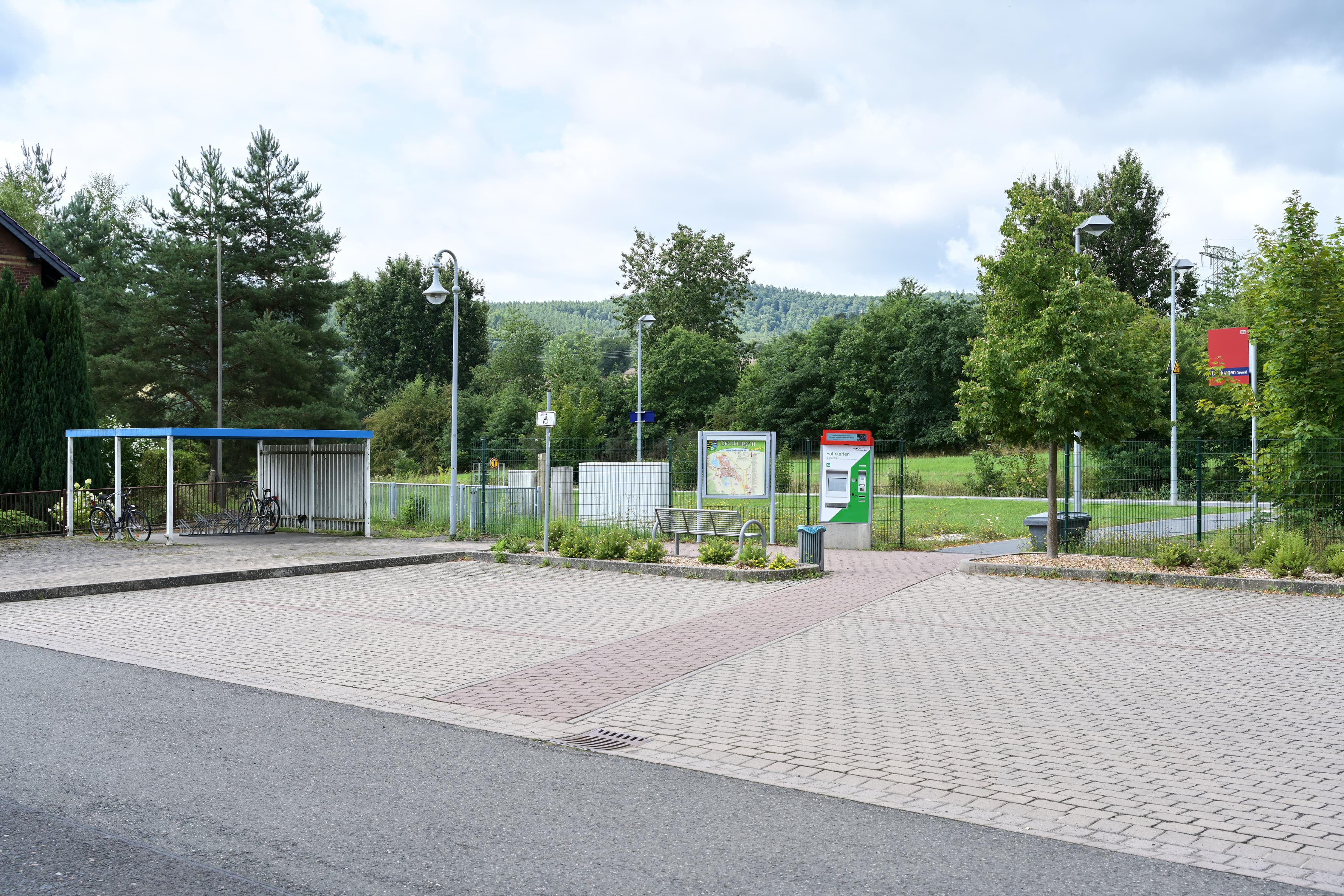 Gesamtblick vom Vorplatz auf die Zuwegung des Haltepunkts. Parkflächen, überdachte Fahrradanlage, Vitrine, Fahrkartenautomat und Zuwegung zum Bahnsteig ist zu erkennen.