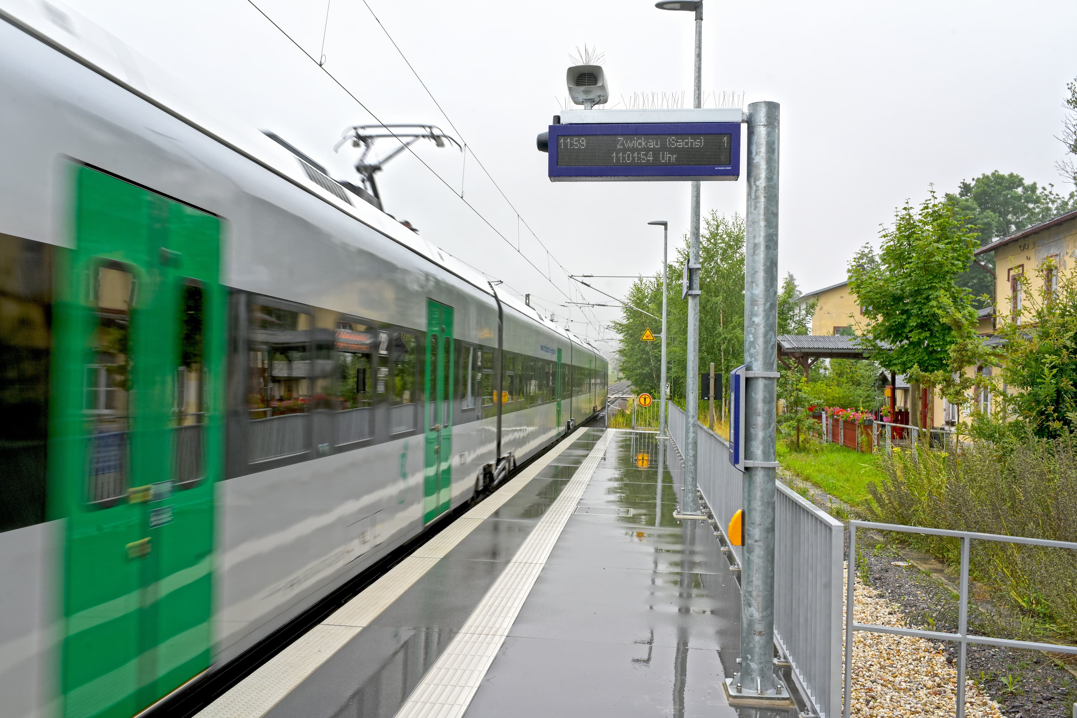 Zu sehen ist ein grau-grüner Zug der am Bahnsteig entlang fährt. Der Bahnsteig ist modern und glänzt vom letzten Regen. 
