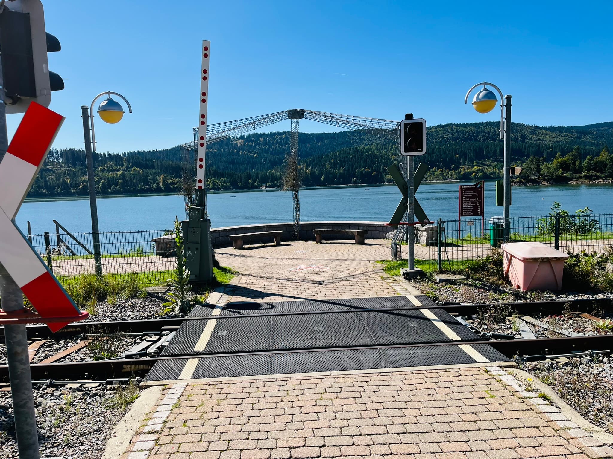 Links eine geöffnete Schranke, die vor dem Bahnübergang steht. In der Mitte zwei Sitzbänke die einen perfekten Blick zum Schluchsee ermöglichen. Hinter dem Schluchsee ist die Natur des Schwarzwaldes zu begutachten. 