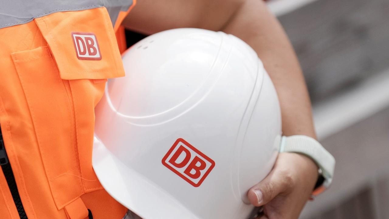 A DB employee in safety vest holding a safety helmet with DB logo in his hands.