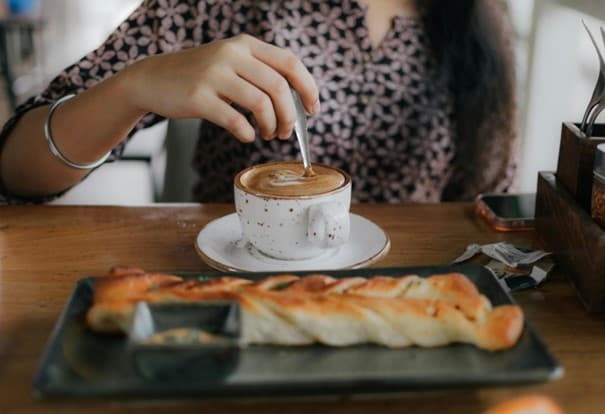 Eine Frau rührt in ihrem Cappuccino, vor ihr steht ein Gebäck auf dem Tisch.