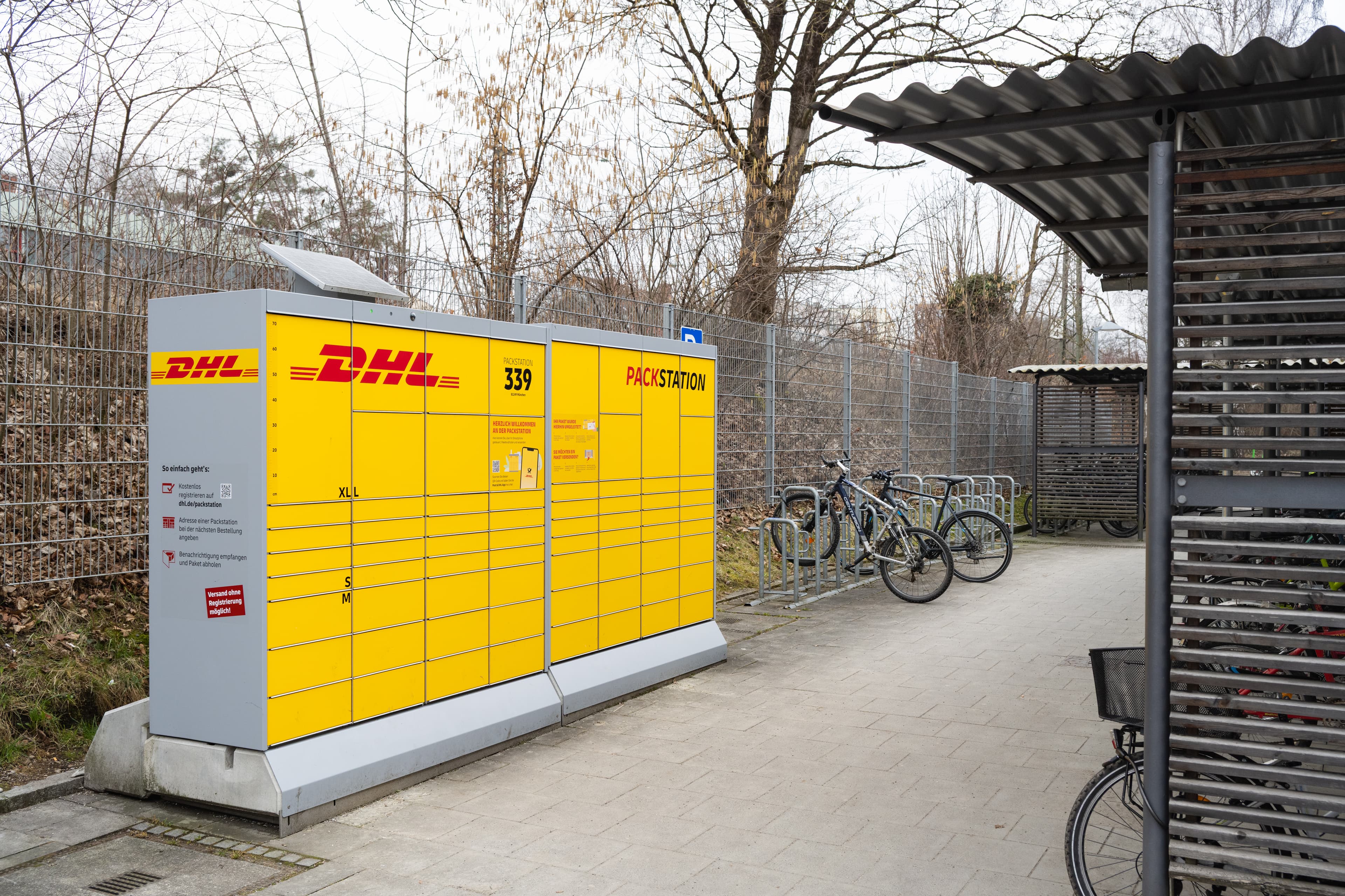 Blick auf eine DHL-Packstation. Rechts daneben sind in einer Fahrradabstellanlage Fahrräder geparkt.