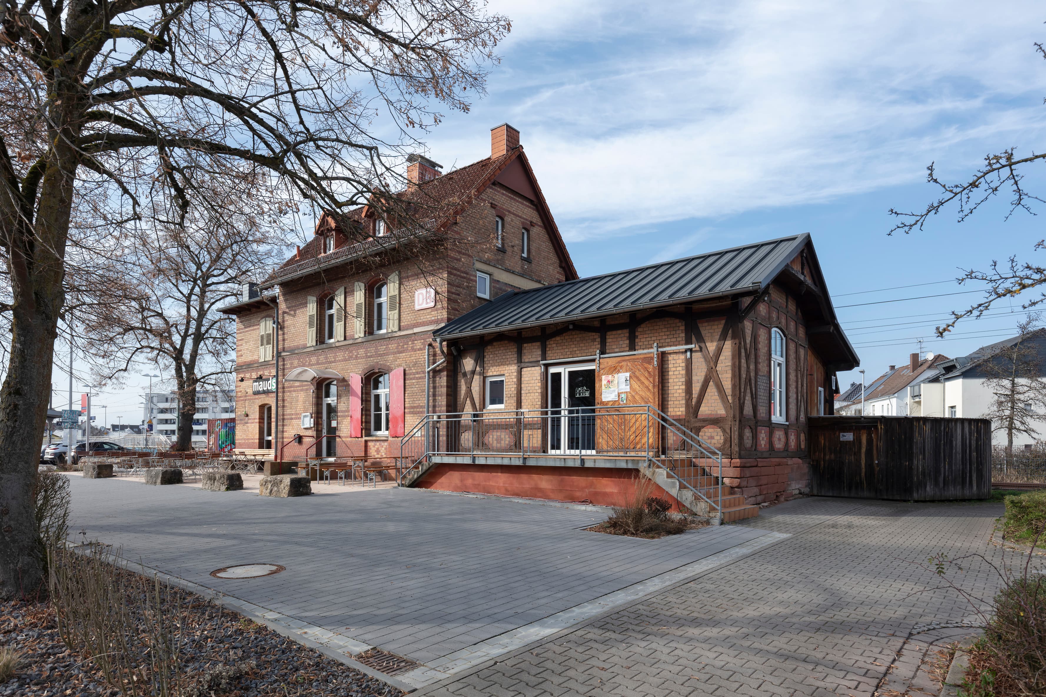 Das blaue Bahnhofsschild mit weißem Schriftzug Rodgau Nieder-Roden neben einem Wetterschutzhaus und einem Metallmülleimer auf einem Bahnsteig.