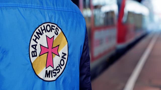 Eine Person am Gleis trägt eine Weste, auf deren Rückseite sich das Logo der Bahnhofsmission befindet.