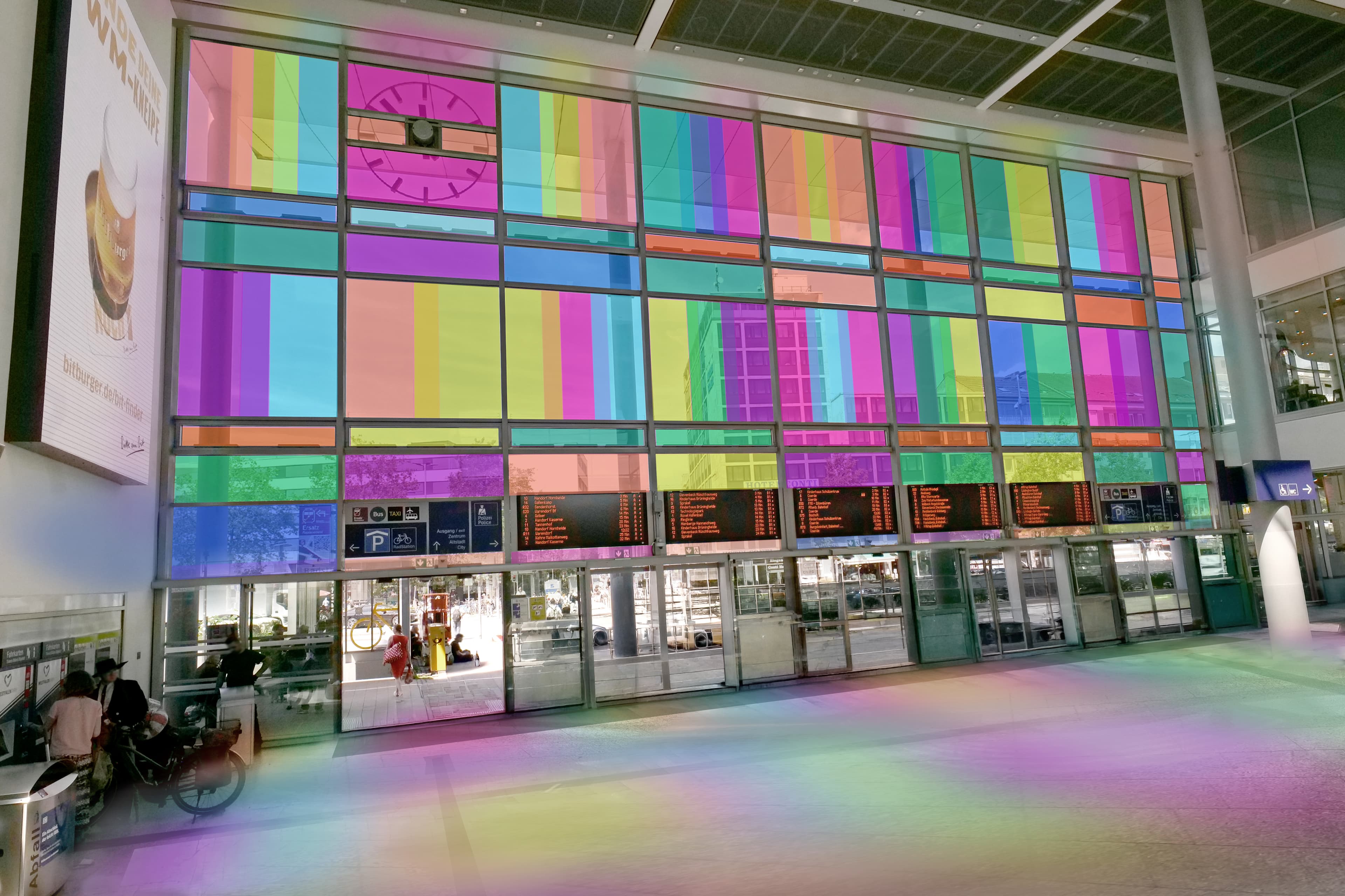 Bahnhofshalle mit bunter Glasfassade und farbigem Licht auf dem Boden.