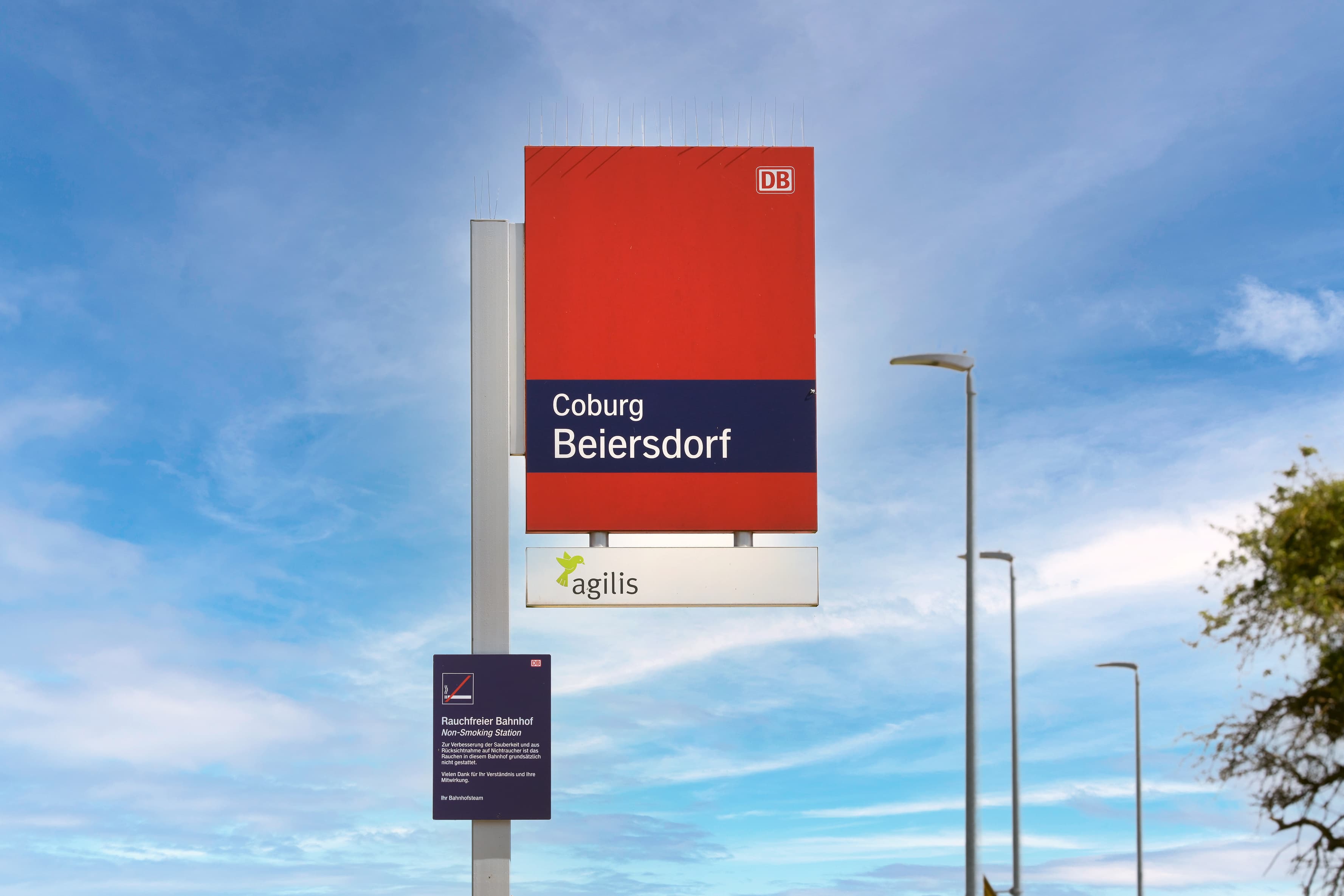 Rotes Bahnhofschild vor blauem Himmel, im Hintergrund hohe Lampen.