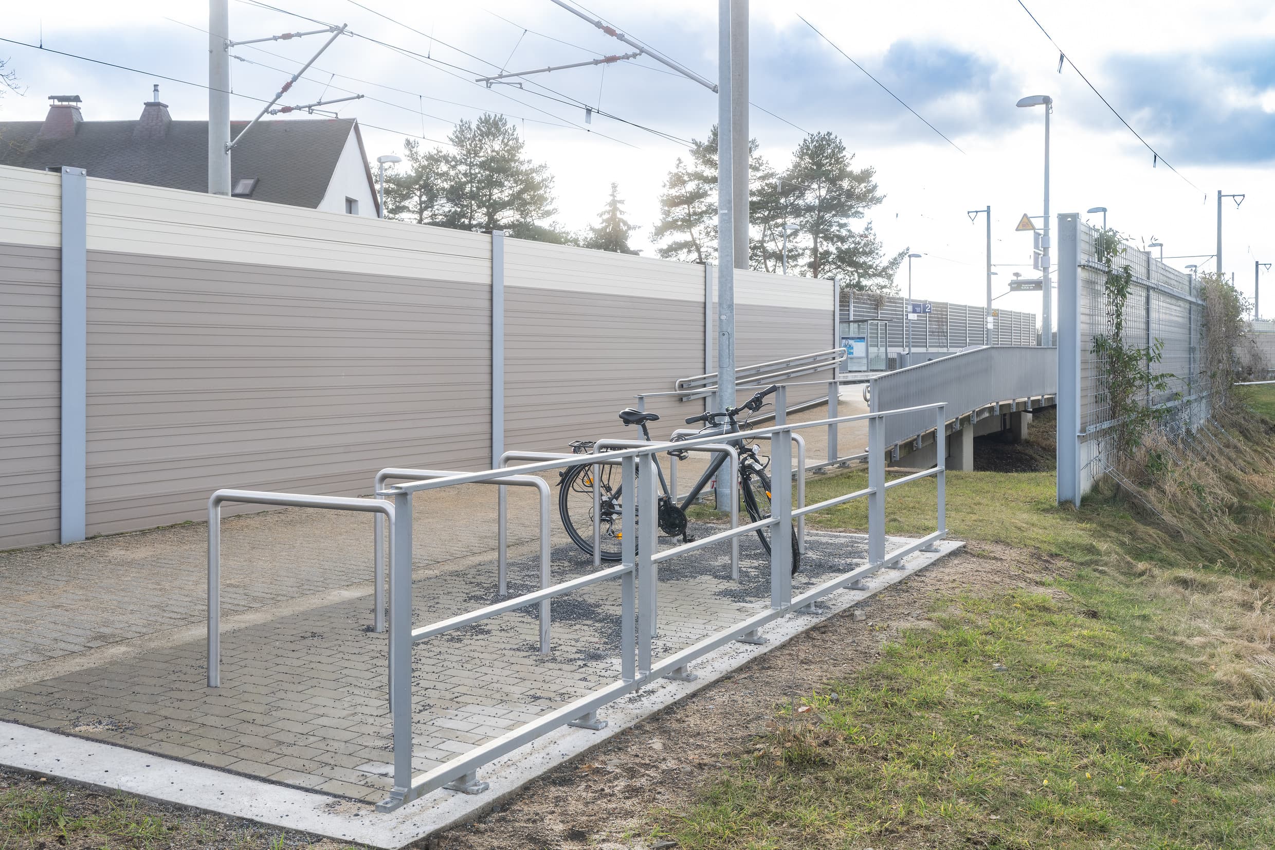 Fünf Einzelbügel für Fahrrädern. An einem Bügel wurde ein Fahrrad abgestellt. Dahinter geht der Weg Richtung Bahnsteig weiter. 