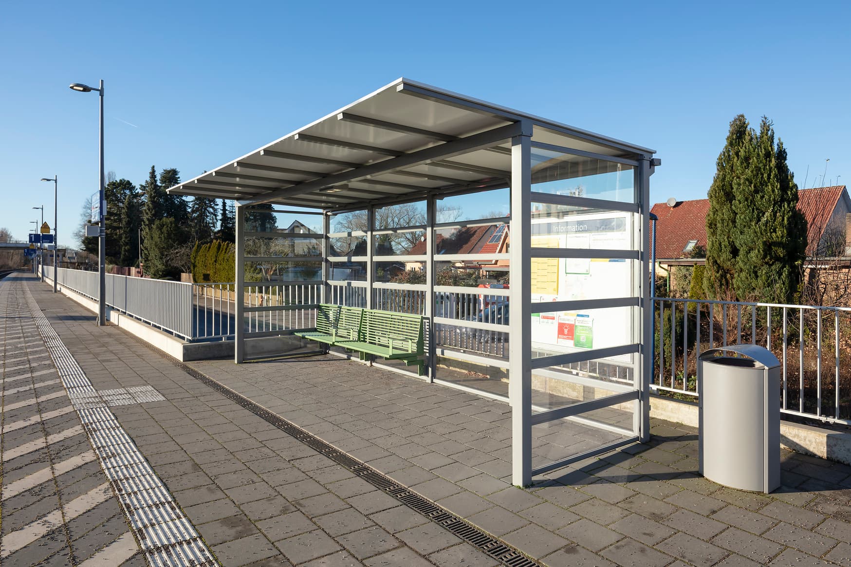 Grüne Sitzbank unter einem gläsernen Wetterschutzhäuschen. 
