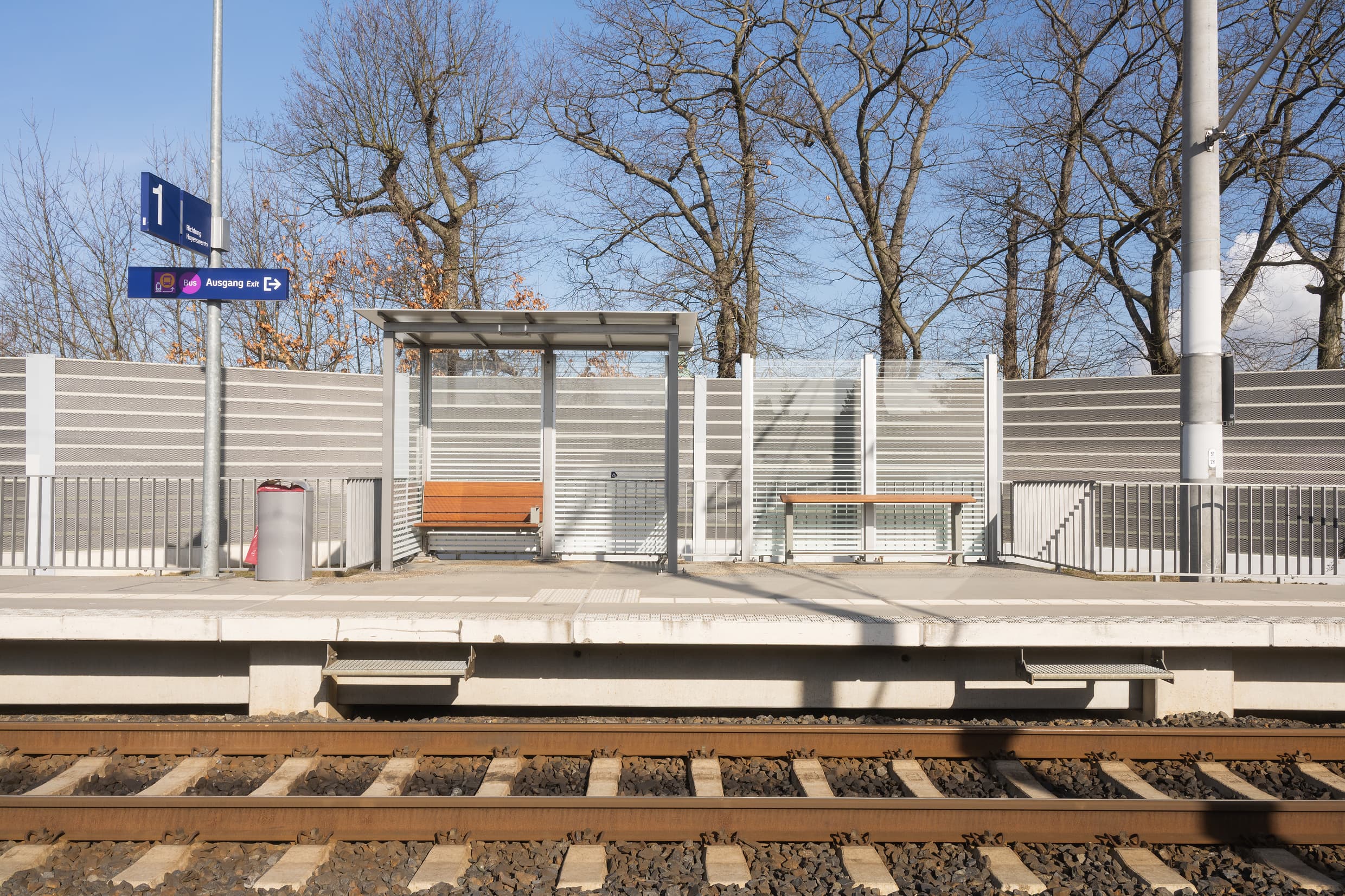 Volltransparentes Wetterschutzhaus mit neuer Holzbank, rechts daneben neuer zusätzlicher Anlehner aus Holz. Eine Vogelsilhouette am Geländer ist ebenfalls links noch zu sehen.