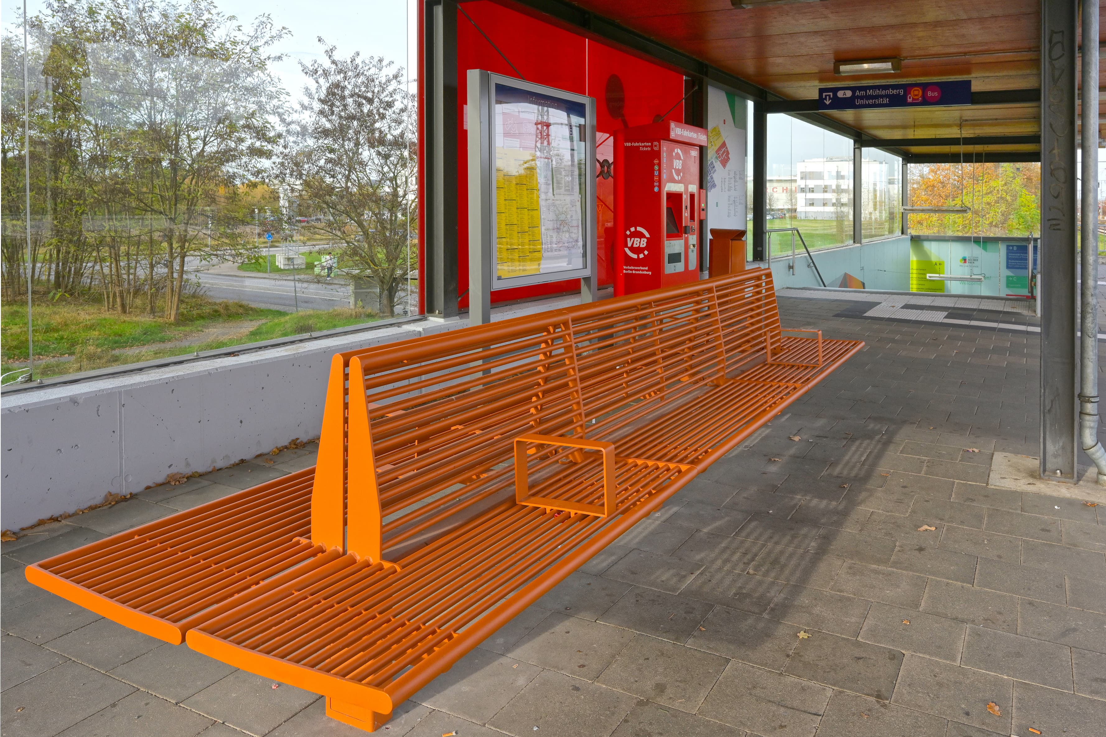 Ein weiterer Blick auf den Wartebereich am Bahnsteig. Im Vordergrund ist eine große, orangene Doppelbank zu sehen. Im Hintergrund die rote Glaswand mit weiterer Bahnsteigausstattung.