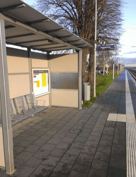 Wetterschutzhäuschen mit Metallpanelen und grauer Sitzbank. Rechts davon ein alter dynamischer Schriftenanzeiger.