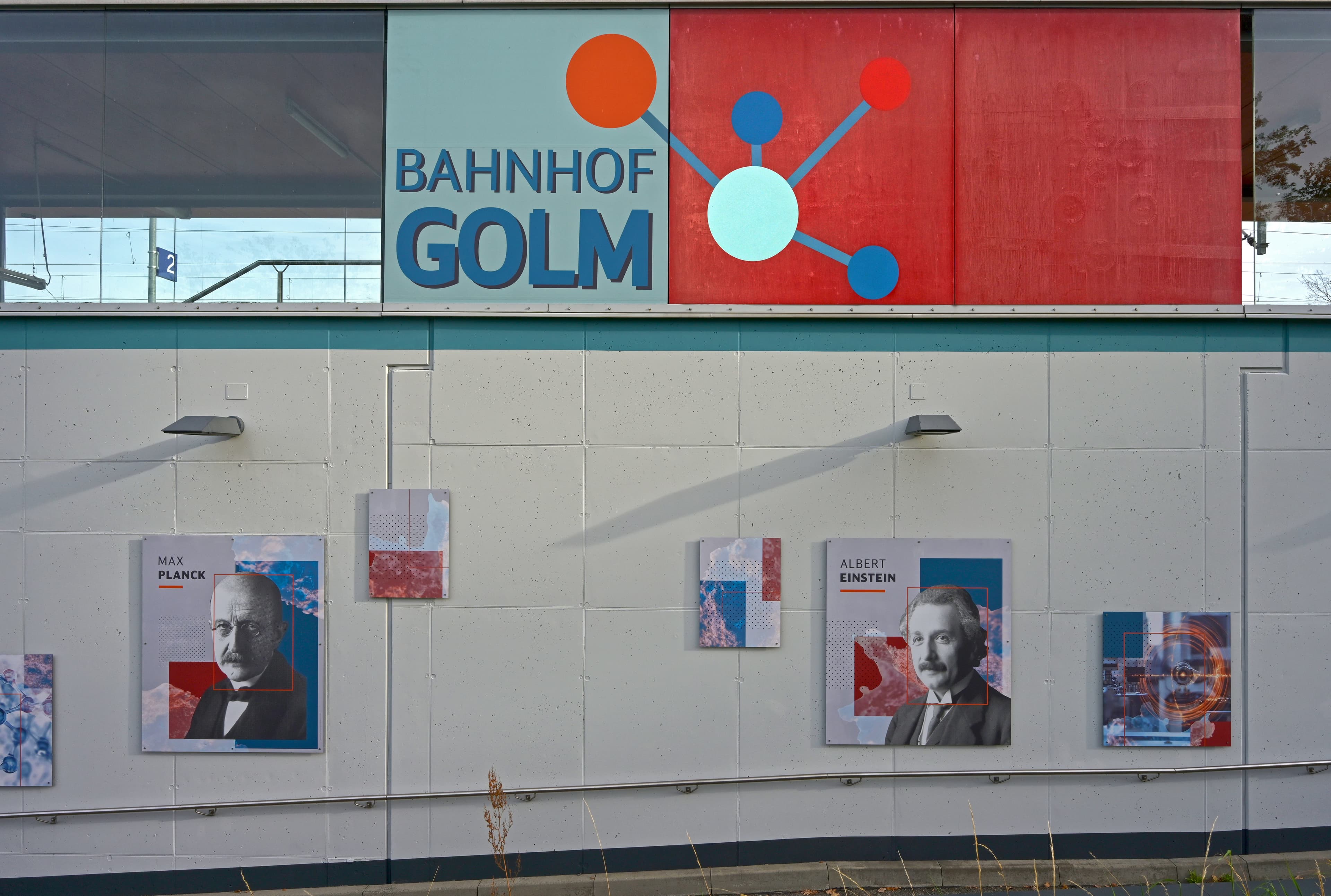 Blick vom Bahnhofsvorplatz. Der Bahnsteig liegt oberhalb des Straßenniveaus und hinter der verzierten, roten Glasfassade. Unterhalb ist ein Teil der Rampe zu sehen - an der Wand des Bahnsteig hängen Porträts von Wissenschaftler:innen.