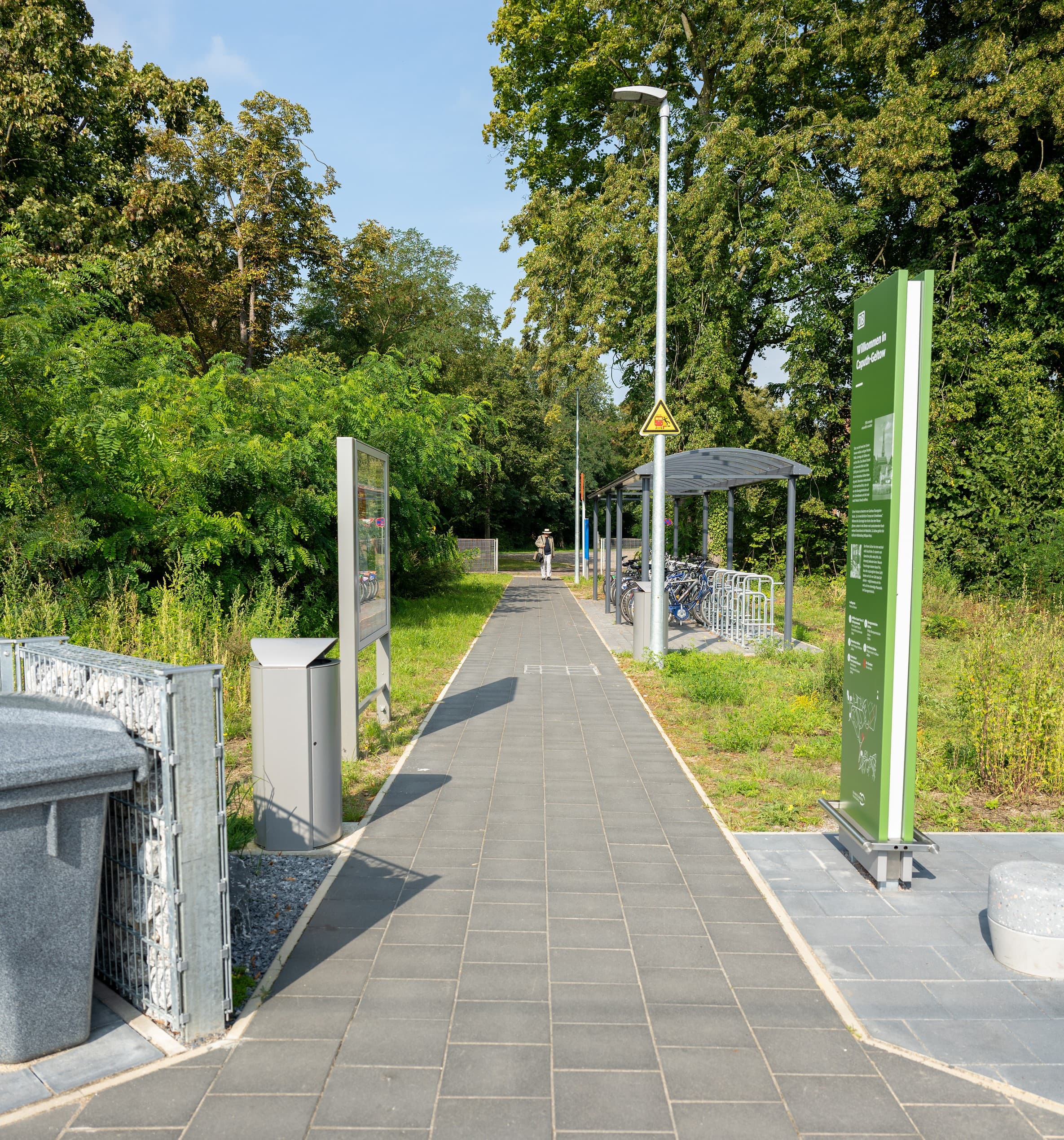 Ein gepflasterter Weg, links ein Mülleimer und eine Vitrine, rechts eine Infostele und ein Fahrradparkhaus. 