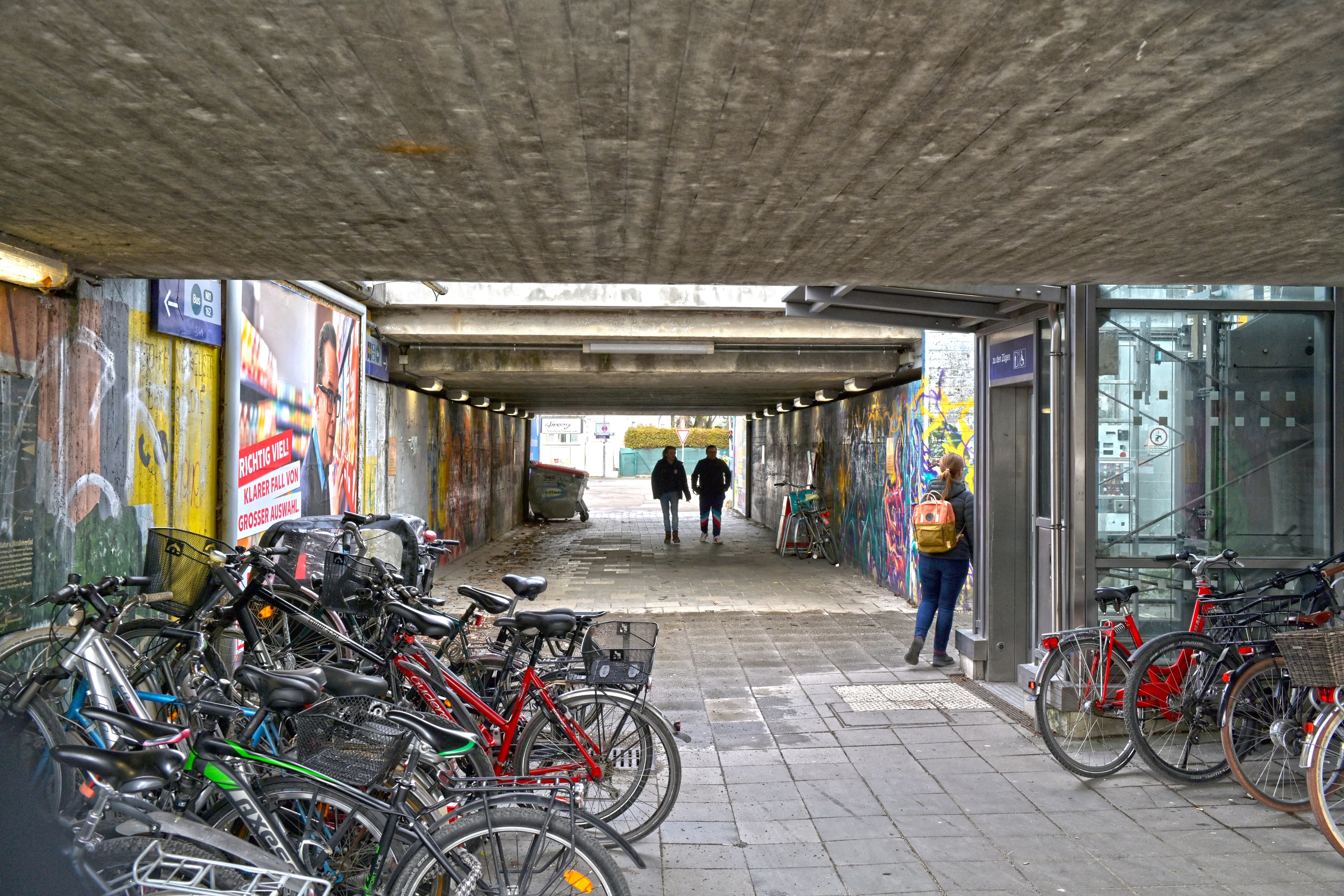 Alte Ansicht der Personenunterführung mit dunklen Wänden und veralteter Gestaltung. Im Vordergrund links und rechts viele abgestellte und abgelegte Fahrräder. Im Zentrum des Bildes gehen Personen durch die dunkle Unterführung.
