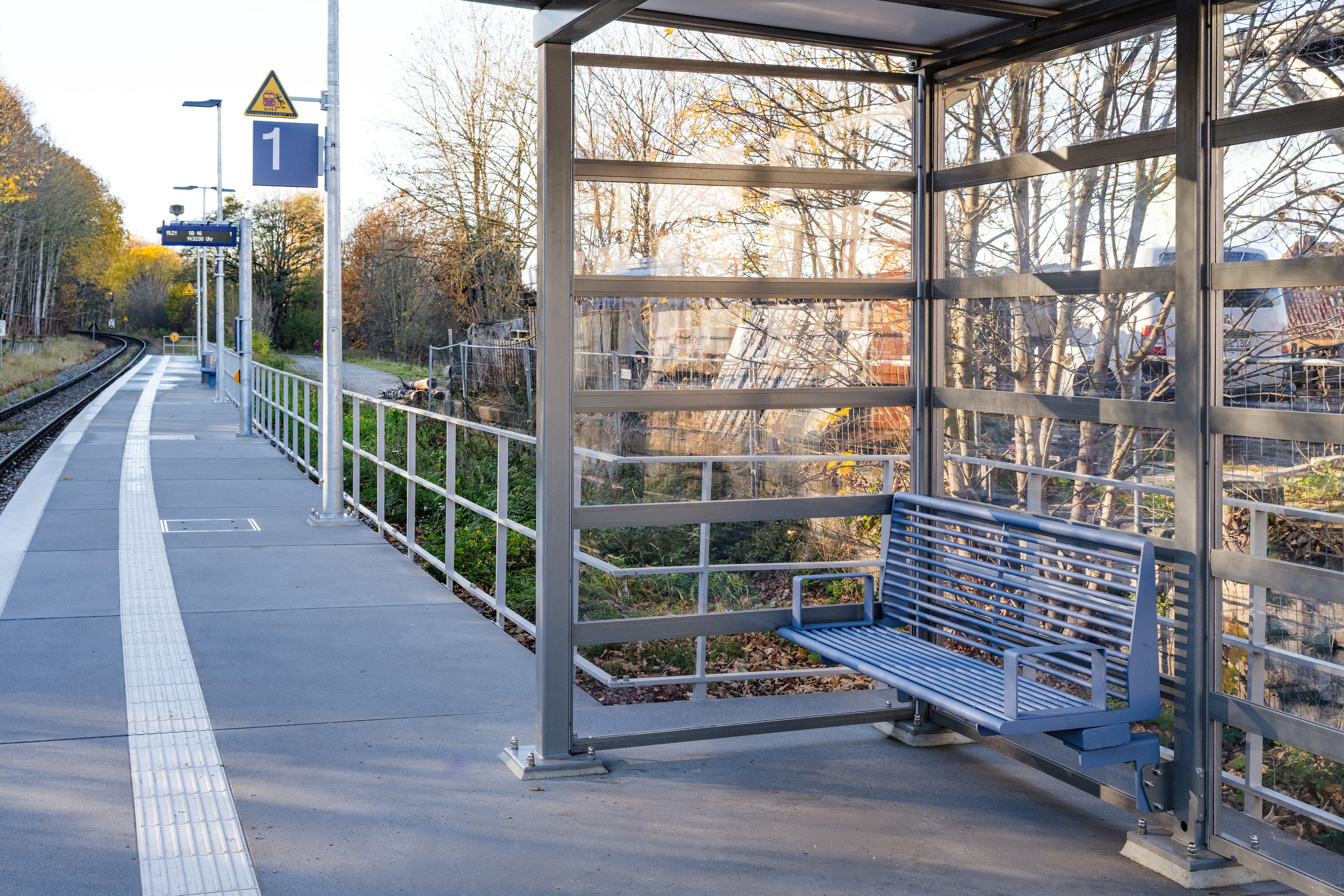 Im Vordergrund sieht man das neue, moderne und volltransparente Wetterschutzhaus mit einer Bank aus blauen Stahlrohren. Im Hintergrund sieht man den umgebauten Bahnsteig und den digitalen Anzeiger.