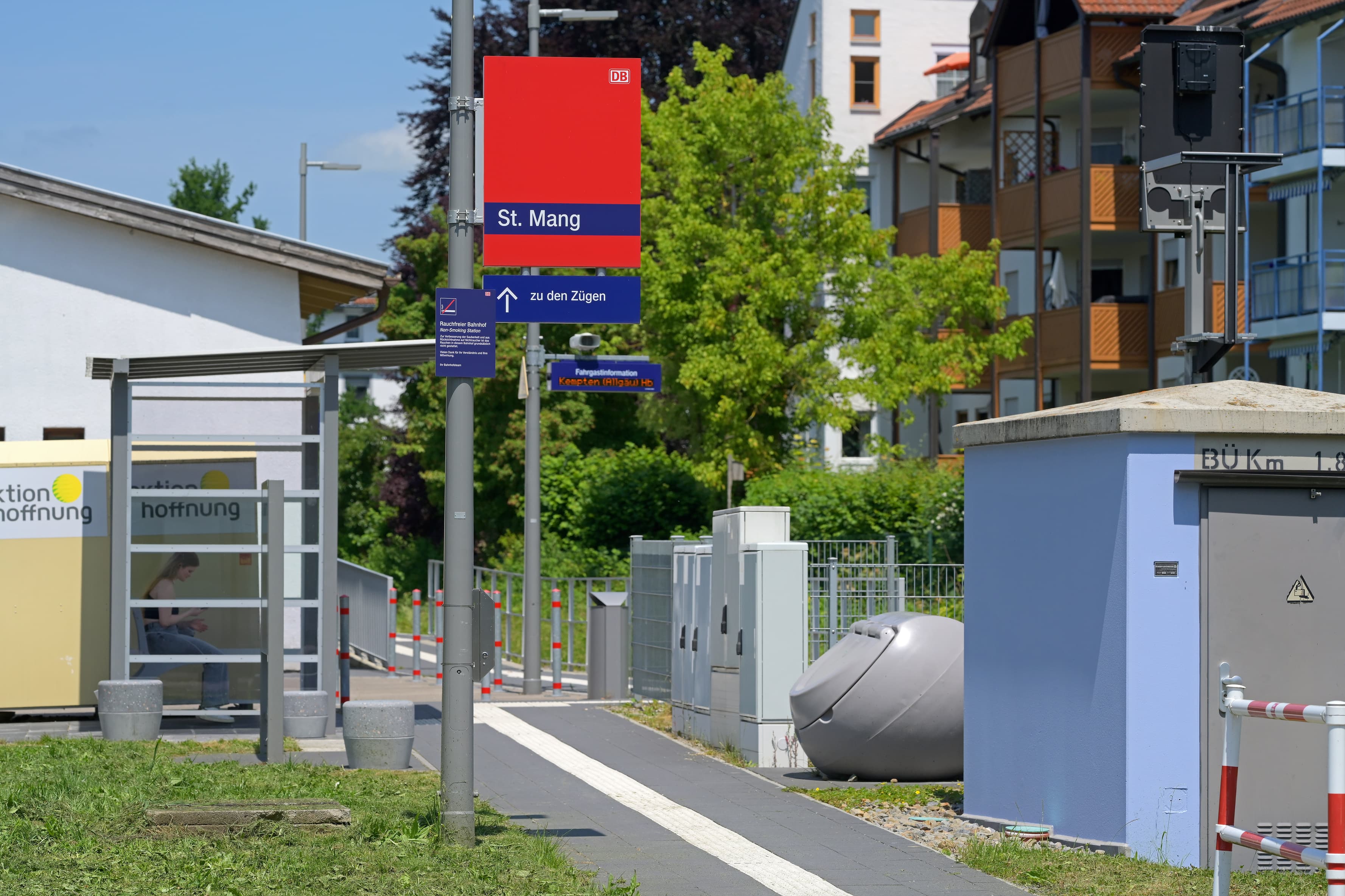 Rechts ein Technikhüschen. Links ein transparentes Wetterschutzhaus. Dazwischen der Weg zum Bahnsteig mit taktilem Leitstreifen.