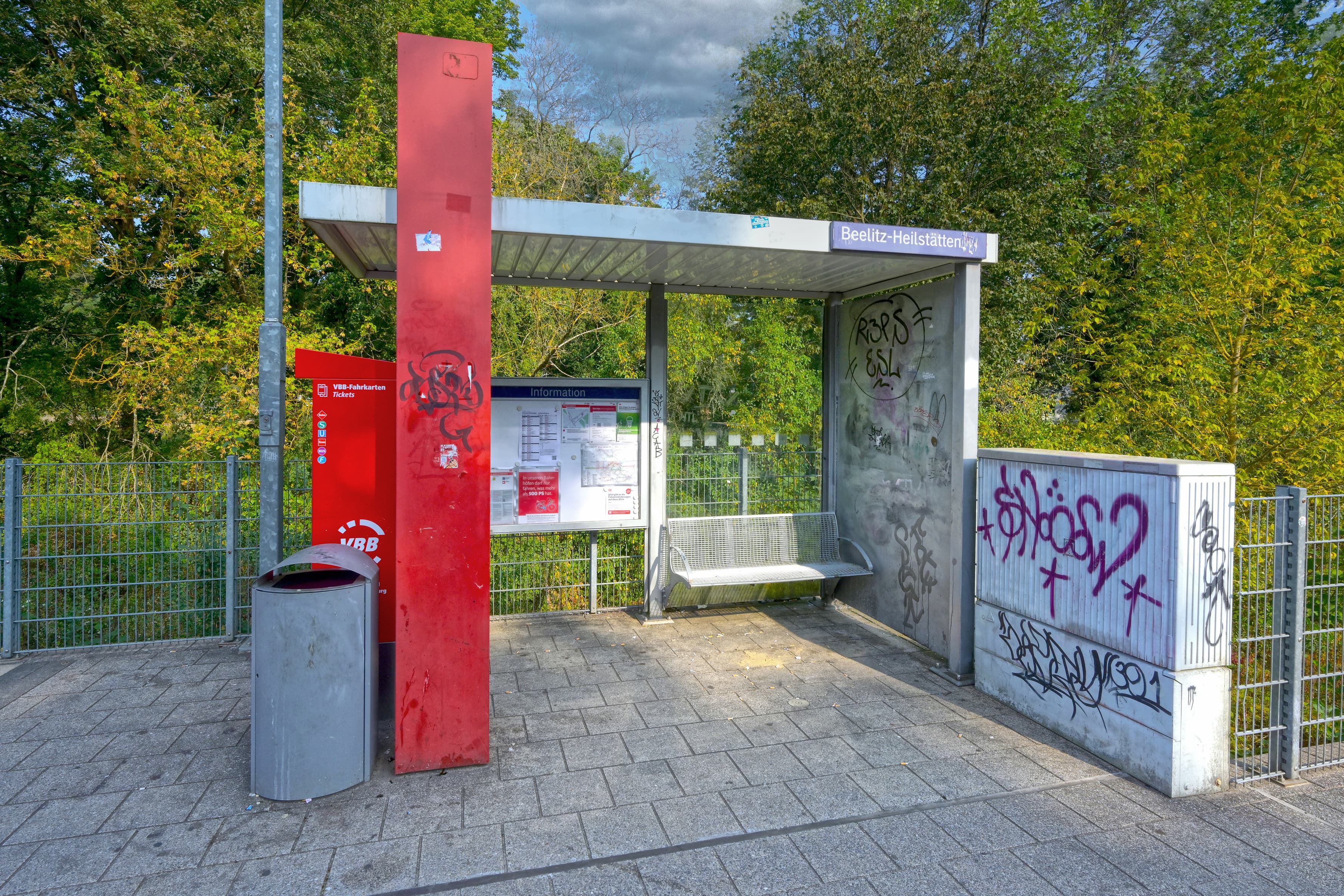 Man sieht ein mit Graffiti beschmiertes Wetterschutzhaus und einen Stromkasten rechts, einen Papierkorb links.