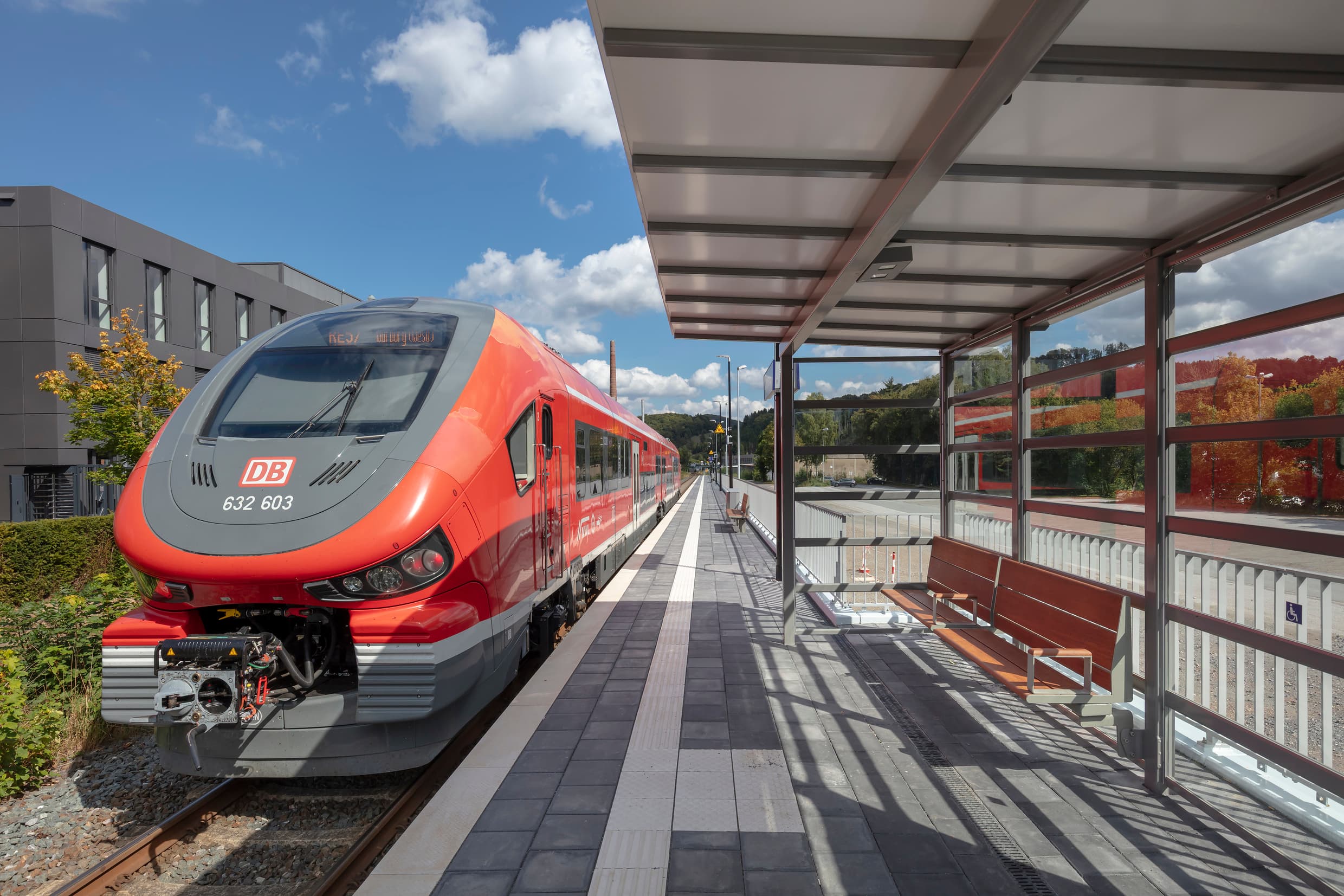Bahnsteig mit WEtterschutzhäuschen und Sitzbank. Ein roter Zug steht am Gleis.
