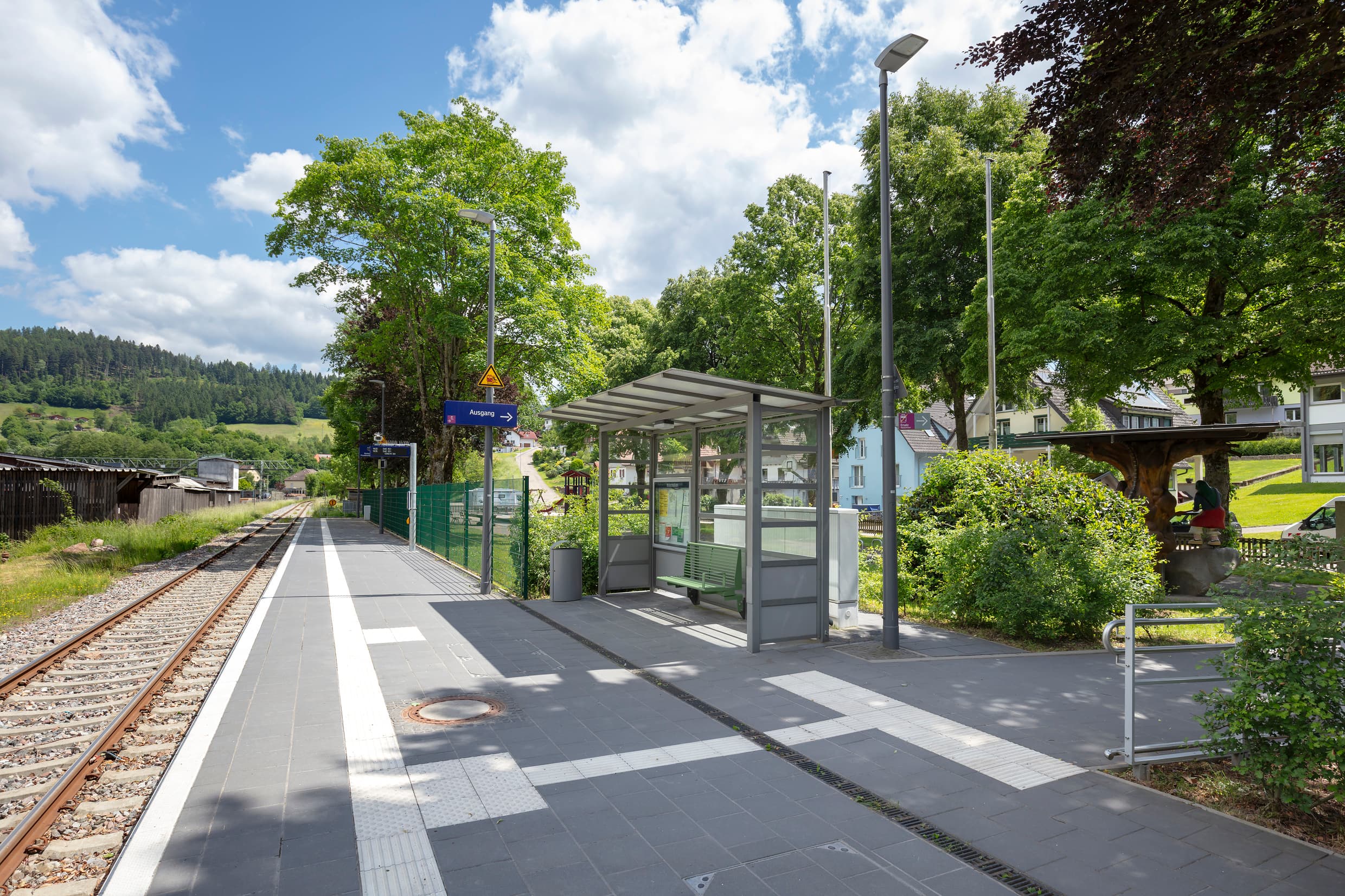 Bahnsteig mit Wetterschutzhäuschen, darin eine grüne Sitzbank. Ausgangsschild, im Hintergrund Dynamischer Schriftenanzeiger Plus. Taktiles Leitsystem auf dem Boden.