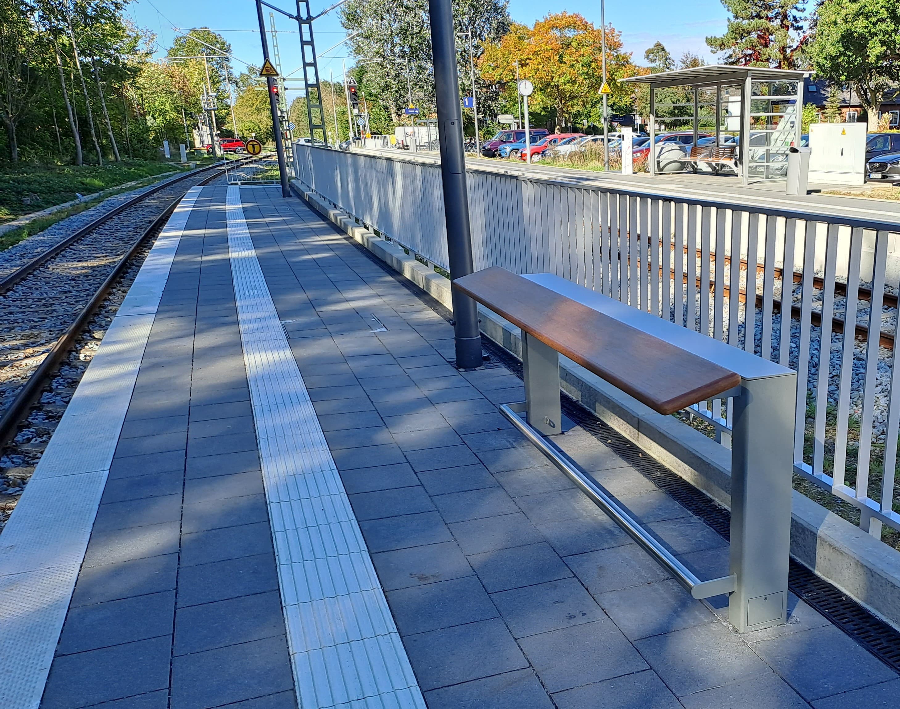 Auf dem gepflasterten Bahnsteig steht ein hüfthoher Anlehner mit Holzoberfläche.