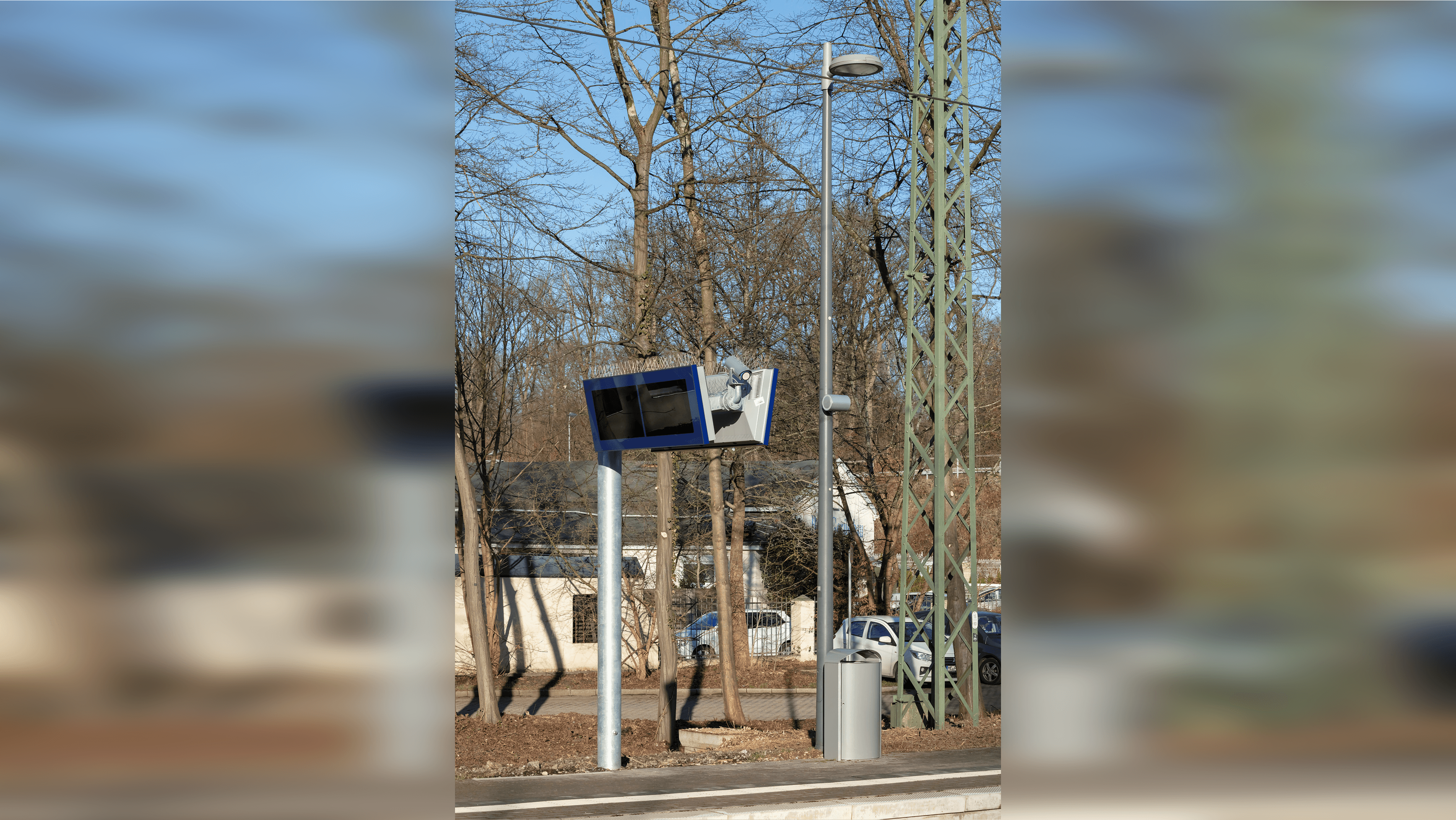 Auf dem Bahnsteig steht ein ZugInoMonitor. Daneben ein Mülleimer aus Metall und eine hohe Laterne an welcher ein Lautsprecher angebracht ist. Im Hintergrund Bäume ohne Blätter.