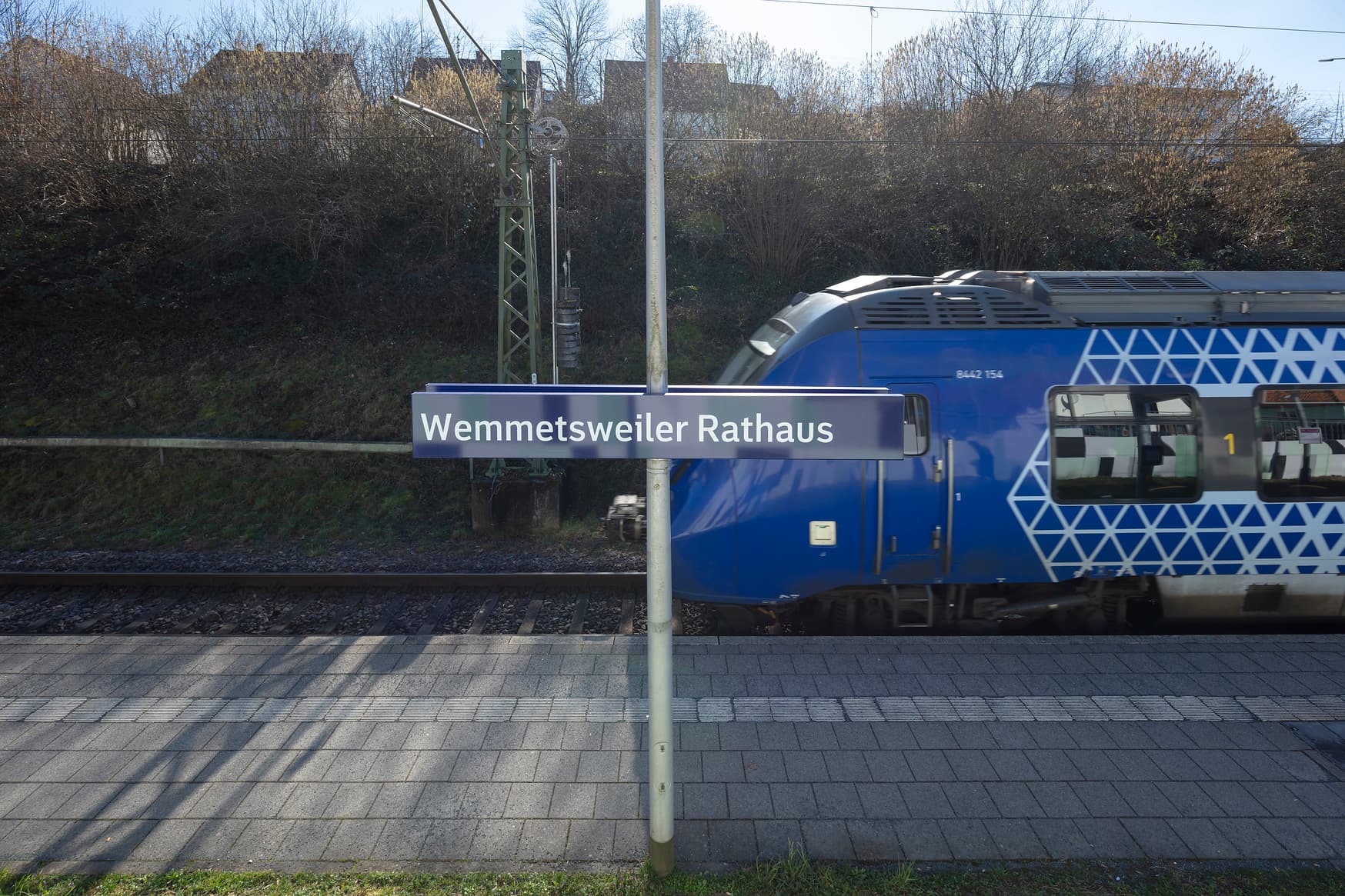 Blick auf Bahnsteig mit taktilem Leitsystem. Im Hintergrund ein einfahrender Zug. Im Vordergrund ein Schild mit der Aufschrift "Wemmetsweiler Rathaus".