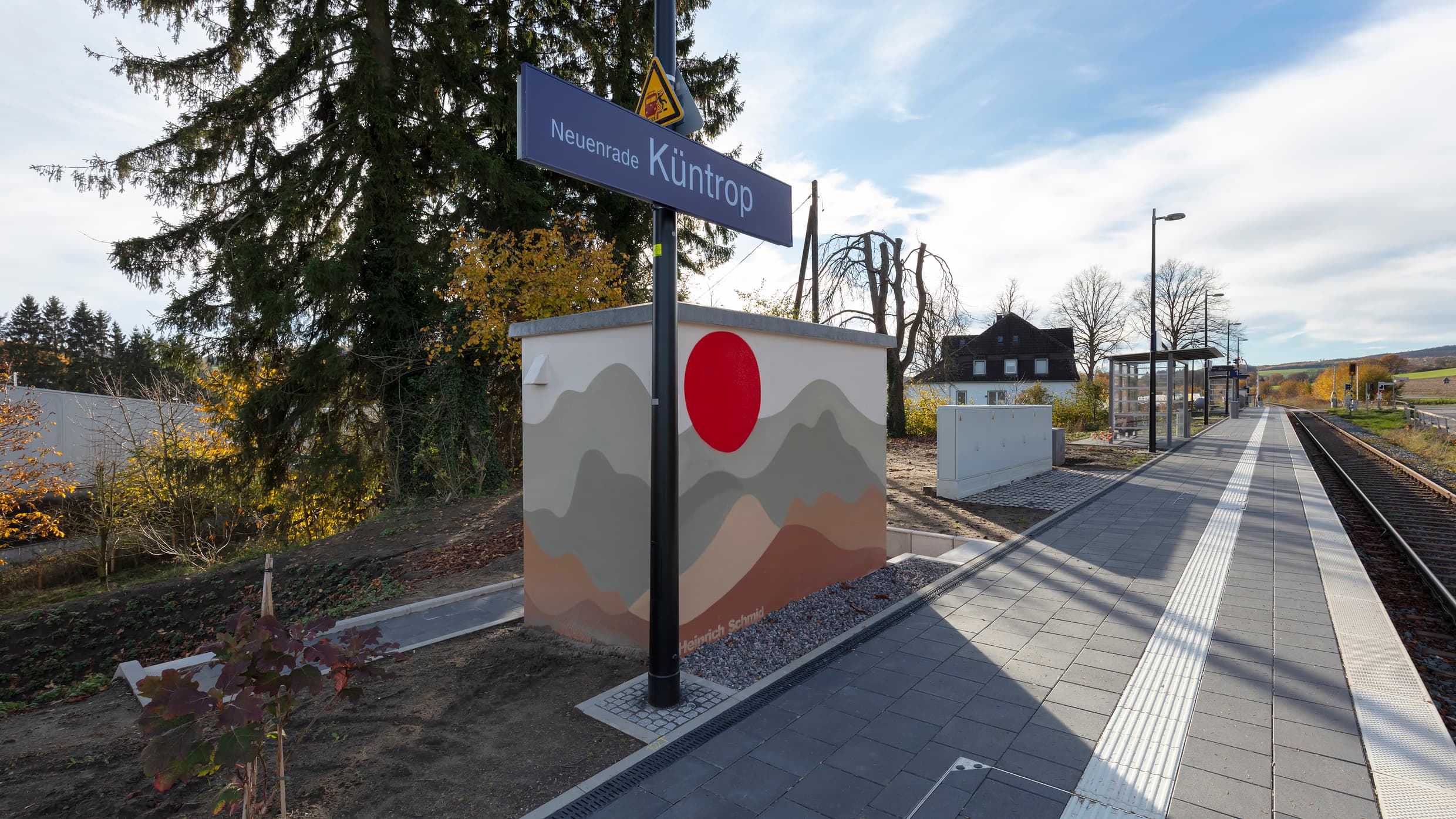 Ein Technikhäuschen am Bahnsteig bemalt mit braun und grau, welches an eine Berglandschaft erinnert und einem großen roten Punkt, welches an eine Sonne erinnert.