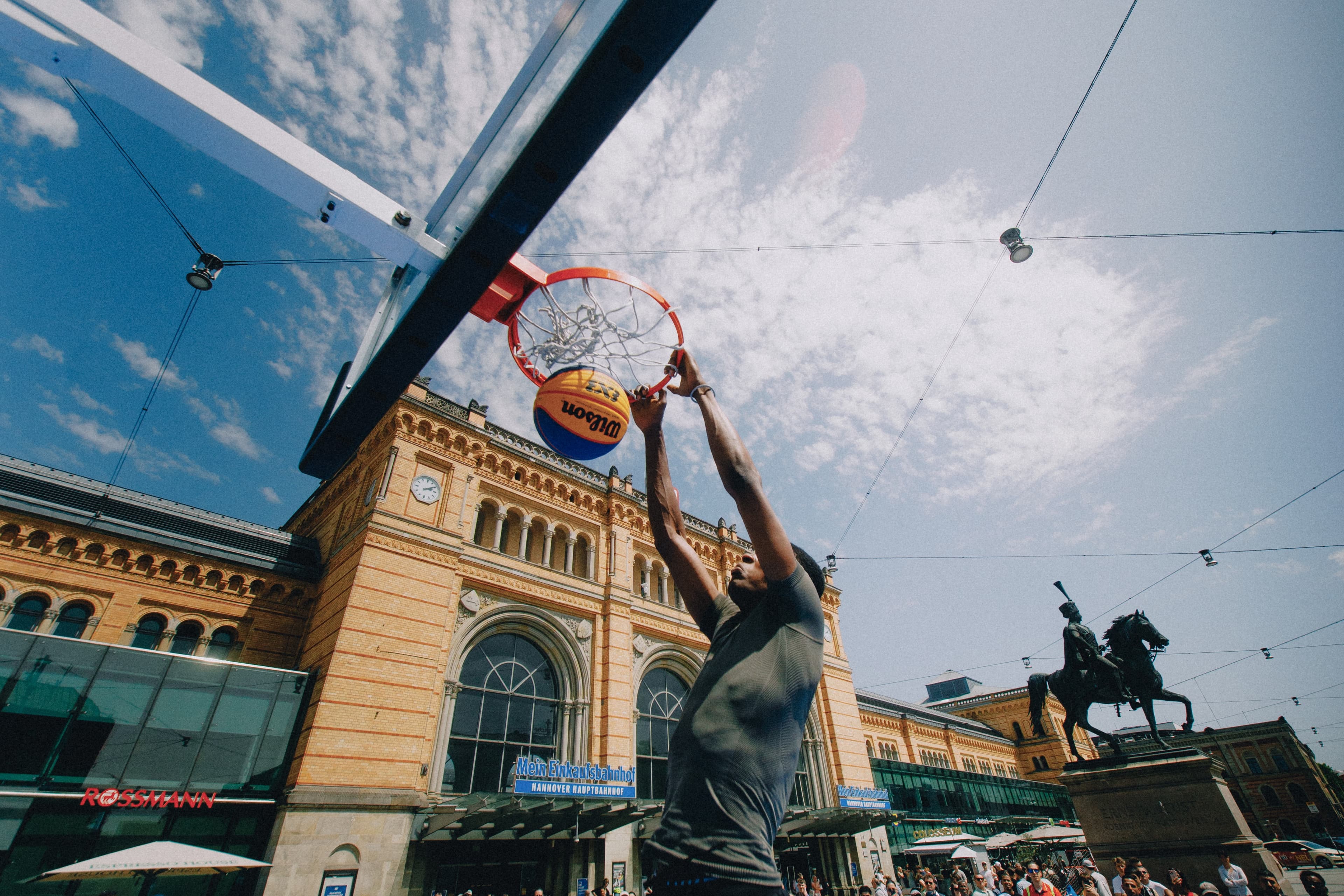 Ein Basketballspieler dunkt vor dem historischen Bahnhofsgebäude des Hannover Hbf im Freien, während Menschen zuschauen und eine Reiterstatue im Hintergrund zu sehen ist.