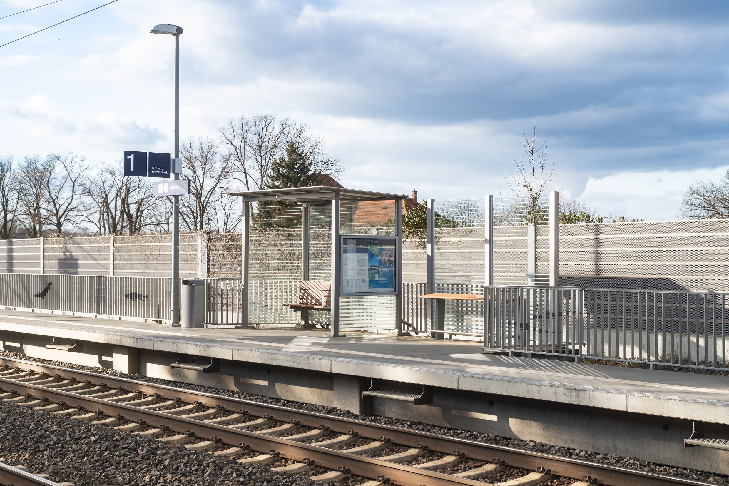 Bahnsteig mit volltransparentem Wetterschutzhaus und Holzbank, rechts daneben steht Anlehner aus Holz, abgegrenzt wird der Bahnsteig durch ein graues Geländer, was mit Fischmotiven gestaltet wurde.
