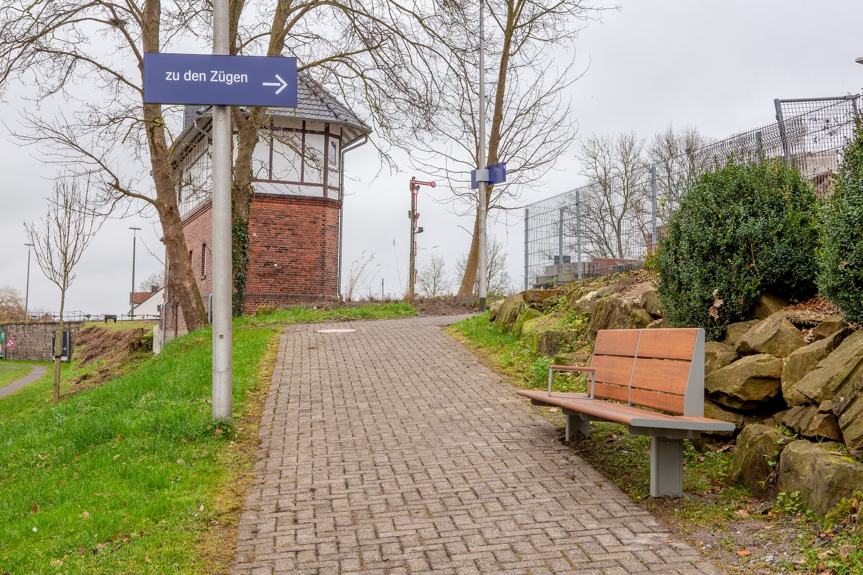 Rampe zum Bahnsteig mit dem blauen Hinweisschild "Zu den Zügen". Am Weg eine Holzbank, im Hintergrund das historische Stellwerk.