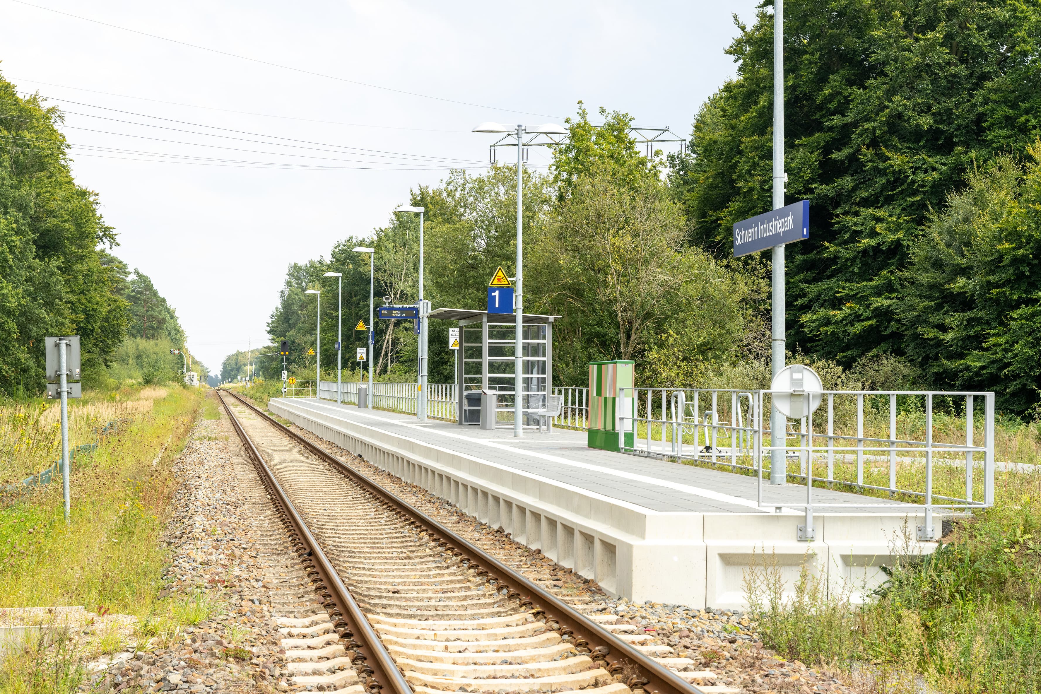 Blick vom örtlichen Bahnübergang aus über den ganzen Bahnsteig. Zu sehen ist ein neuer Bahnsteig mit einladender, heller und sauberer Bahnsteigausstattung (Wetterschutzhaus, Sitzbänke, Anzeigemonitore, etc.)