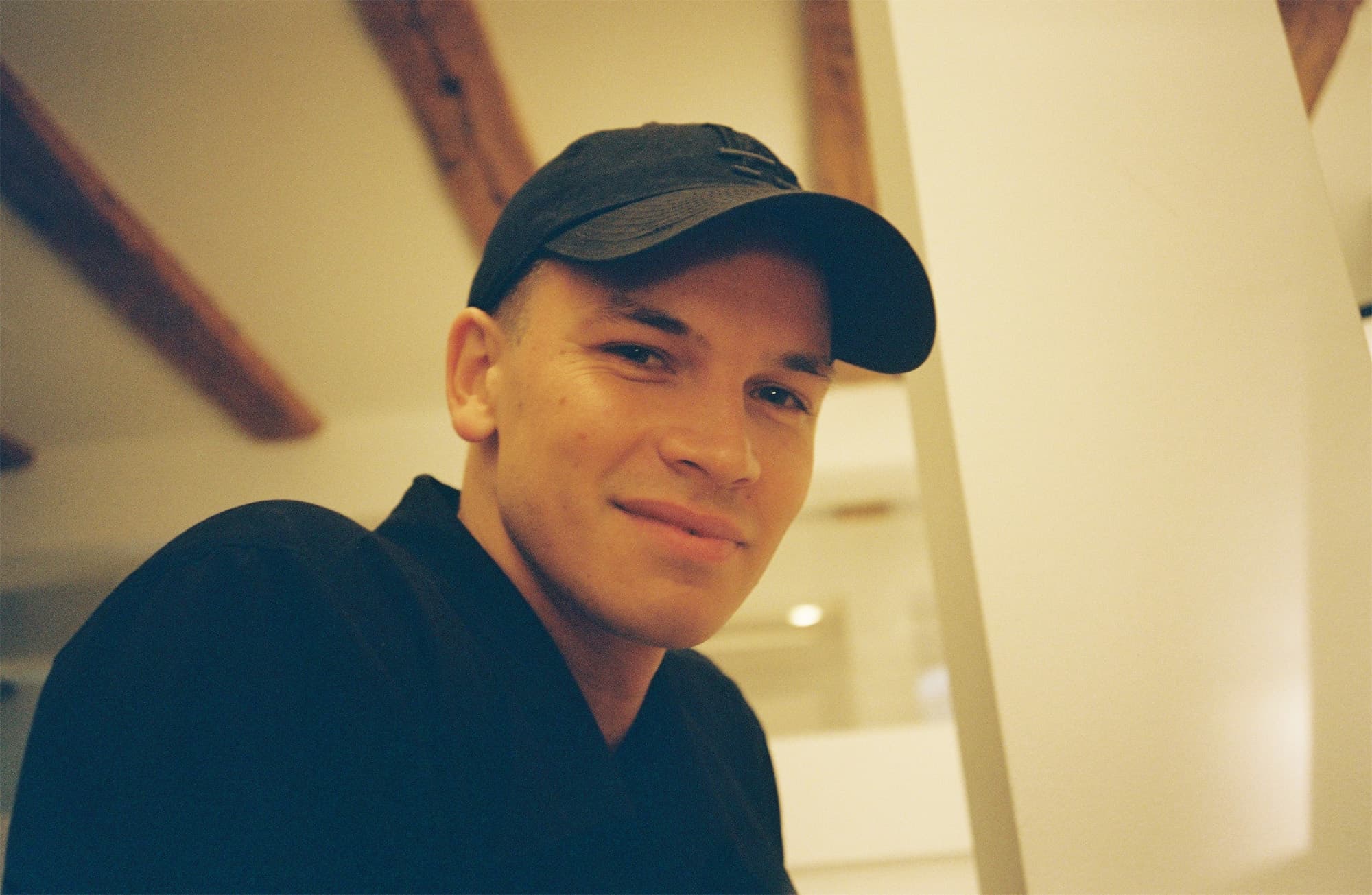 MIK mit schwarzer Kappe und schwarzem Pullover vor einer hellen Wand mit sichtbaren Holzbalken an der Decke