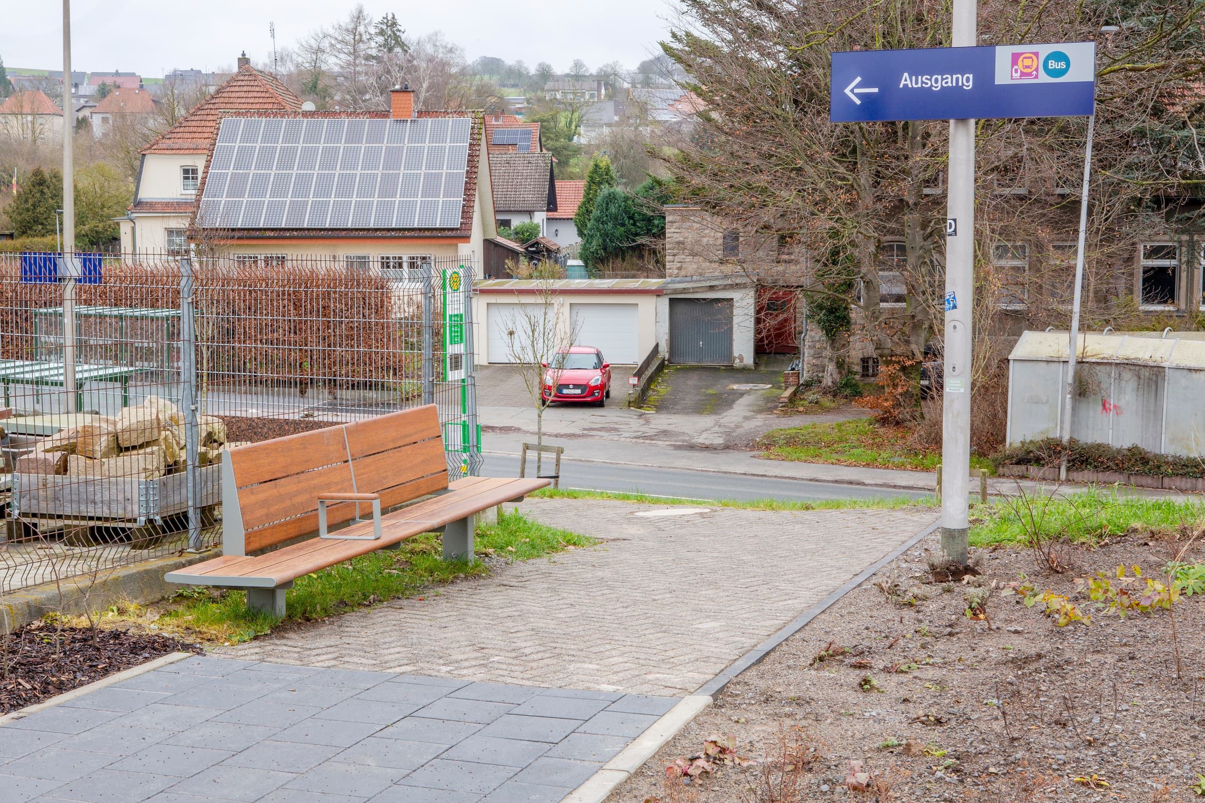 Die Zuwegung vom Bahnhof mit Bambusbank und Schild auf dem Ausgang steht und die Symbolde für Bus und Schienenersatzverkehr abgebildet sind.
